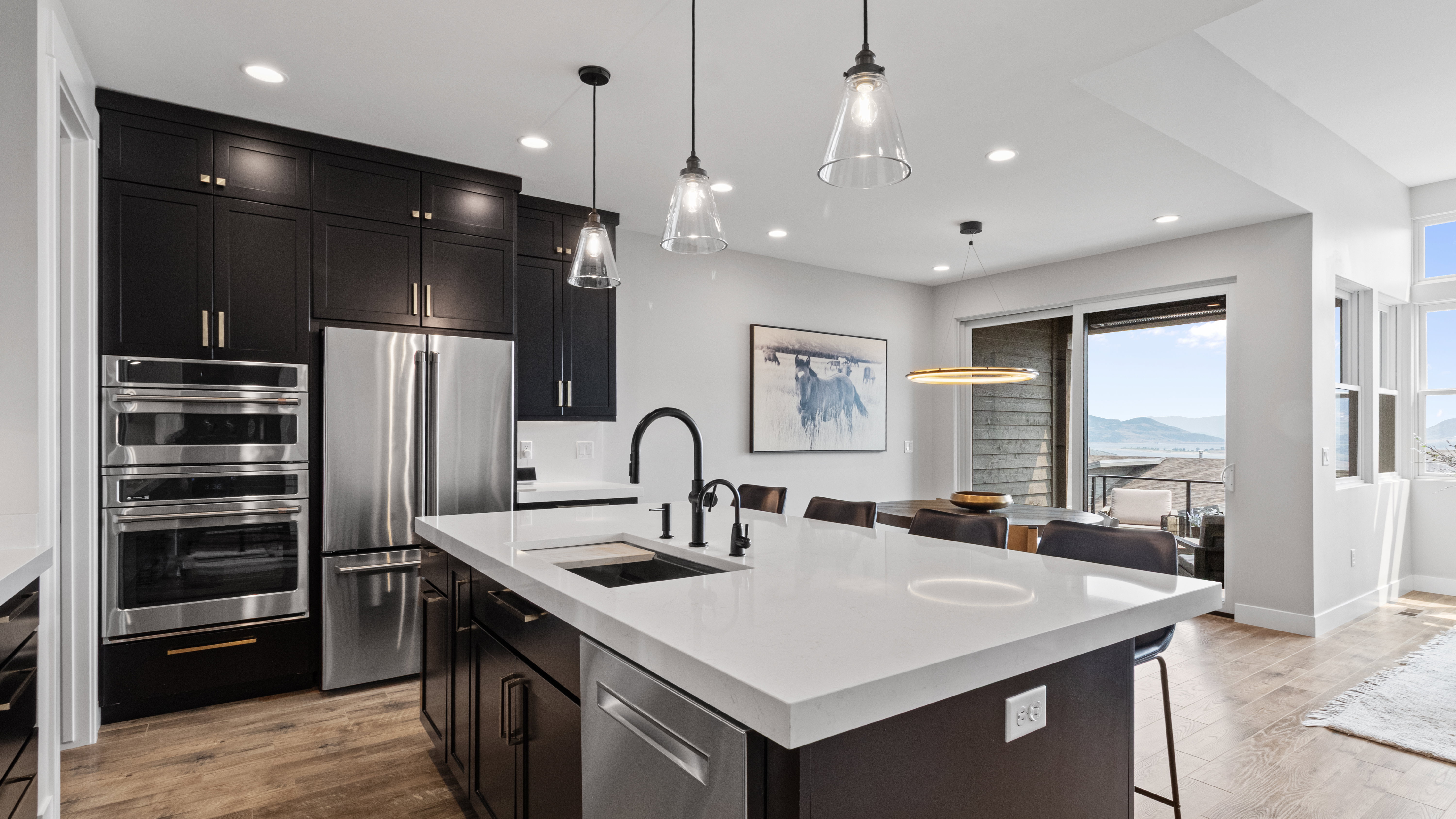 Modern kitchen featuring dark cabinetry, stainless steel appliances, a large center island with a white countertop, wood-finish flooring, and a sliding glass door opening to an outdoor space with mountain scenery