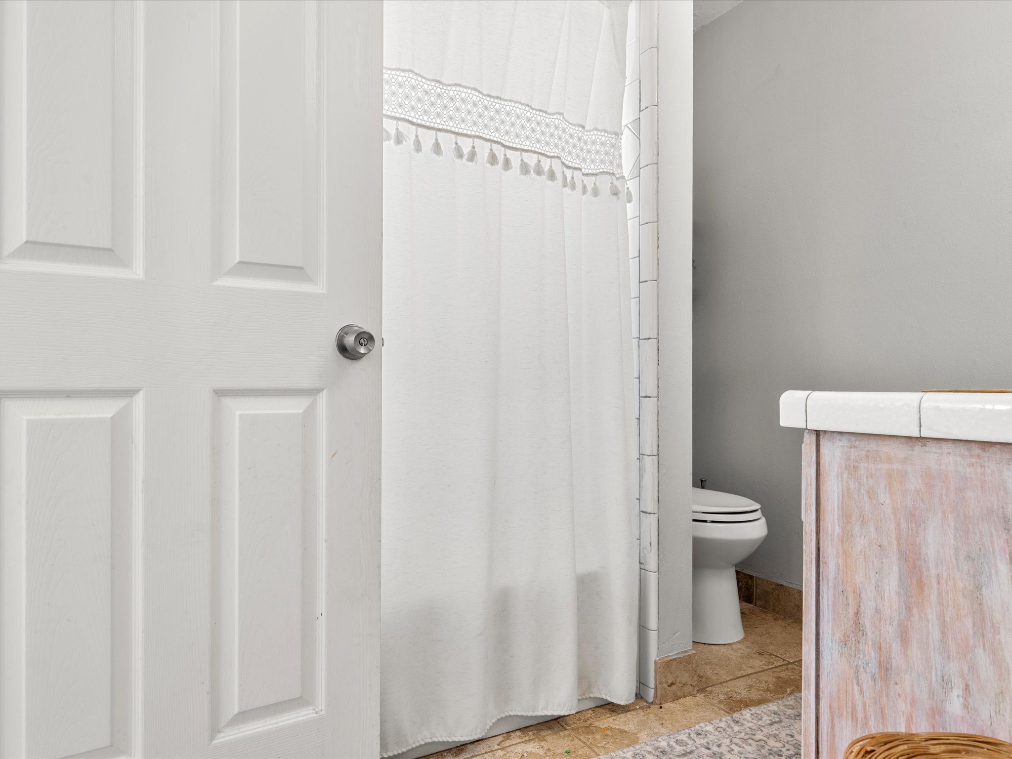 White paneled door opening to a bathroom featuring a tile-top vanity with a wood-finish base, a toilet, and tile flooring