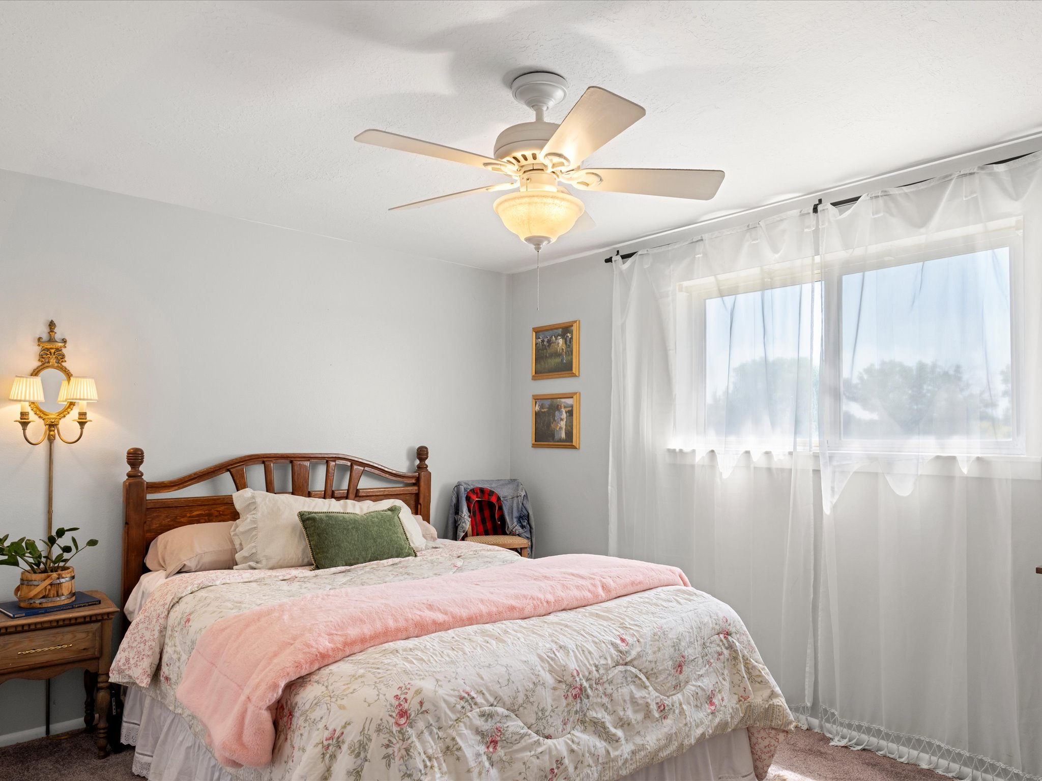 Bedroom featuring a window with sheer curtains, a ceiling fan with integrated lighting, and a wall-mounted sconce