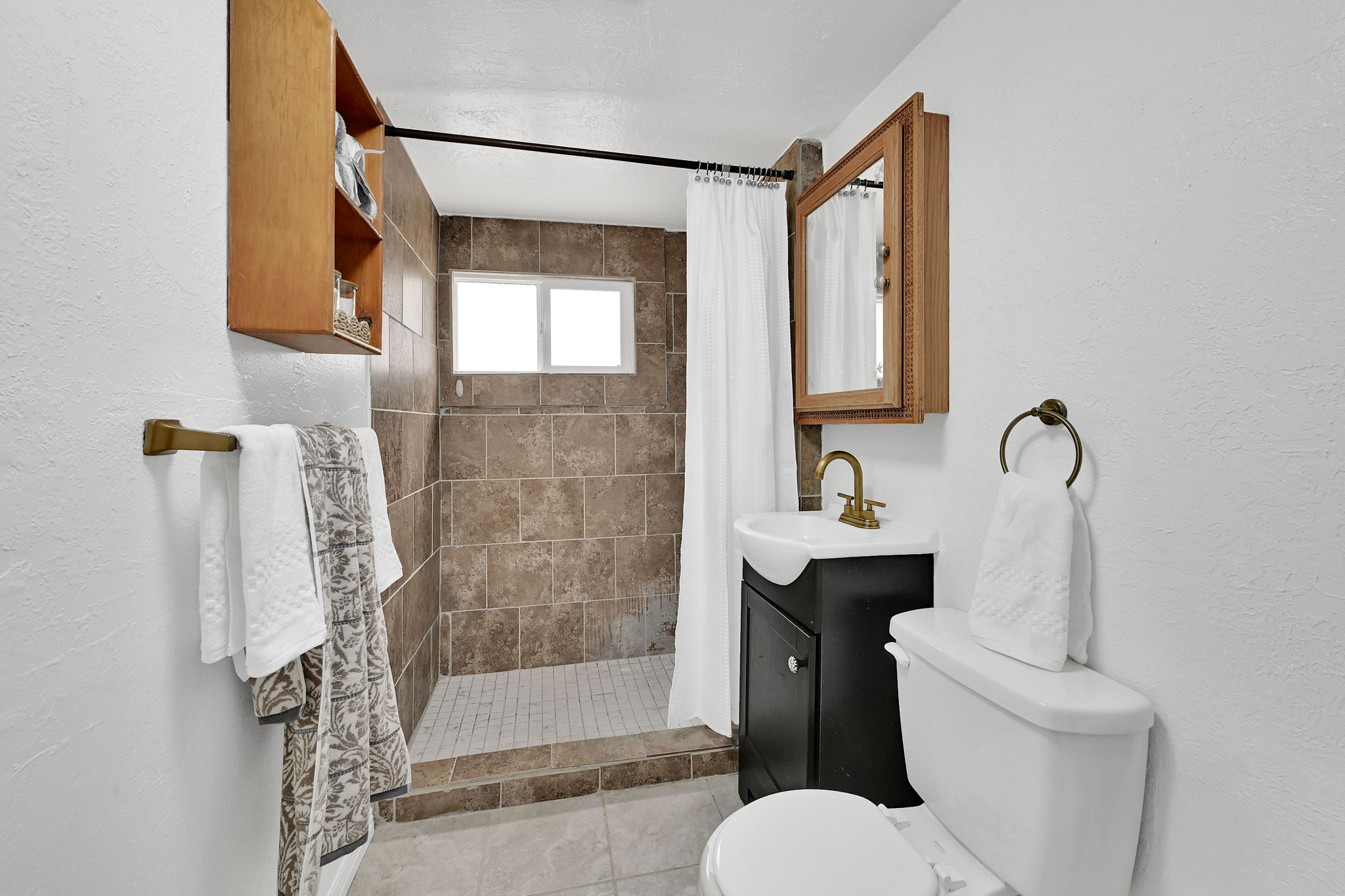 Bathroom featuring a tiled walk-in shower with a window, a dark wood-finish vanity with a white basin sink, a toilet, and a medicine cabinet with a wood-finish frame
