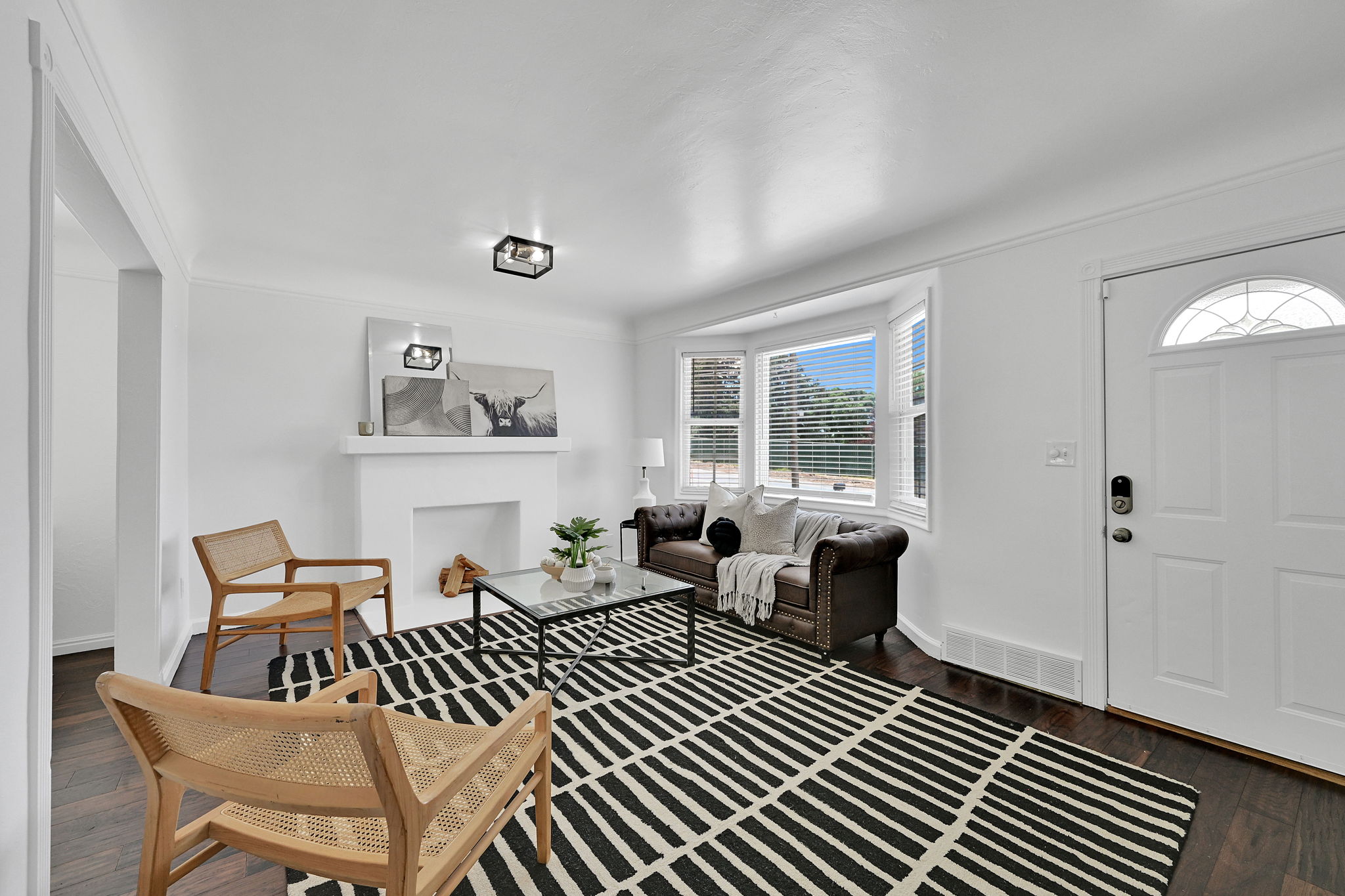 Living area featuring wood-finish flooring, a white-painted fireplace with mantel, and a bay window with blinds