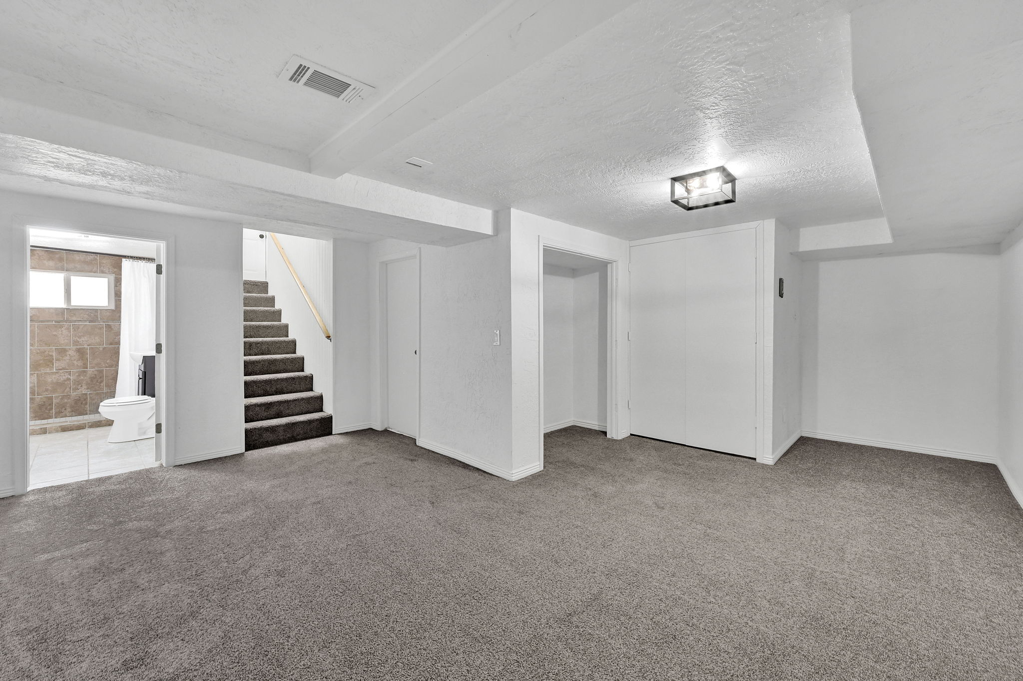 Spacious room featuring gray carpeting, white painted walls, a flush-mount ceiling light, a built-in closet with bifold doors, and an open doorway to a staircase