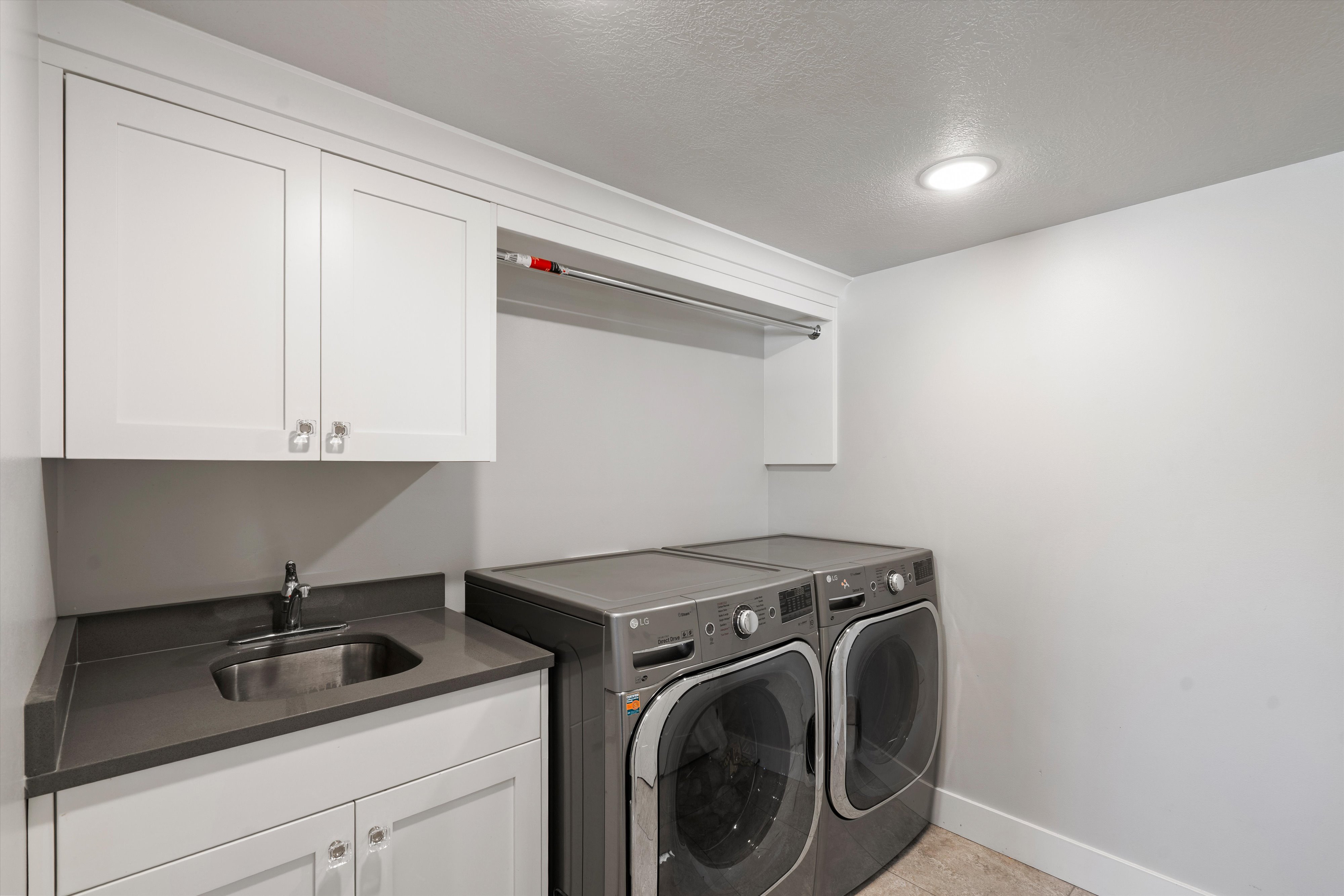 Dedicated laundry area featuring a built-in utility sink with a stainless steel basin, white shaker cabinetry, dark countertop, and integrated overhead lighting
