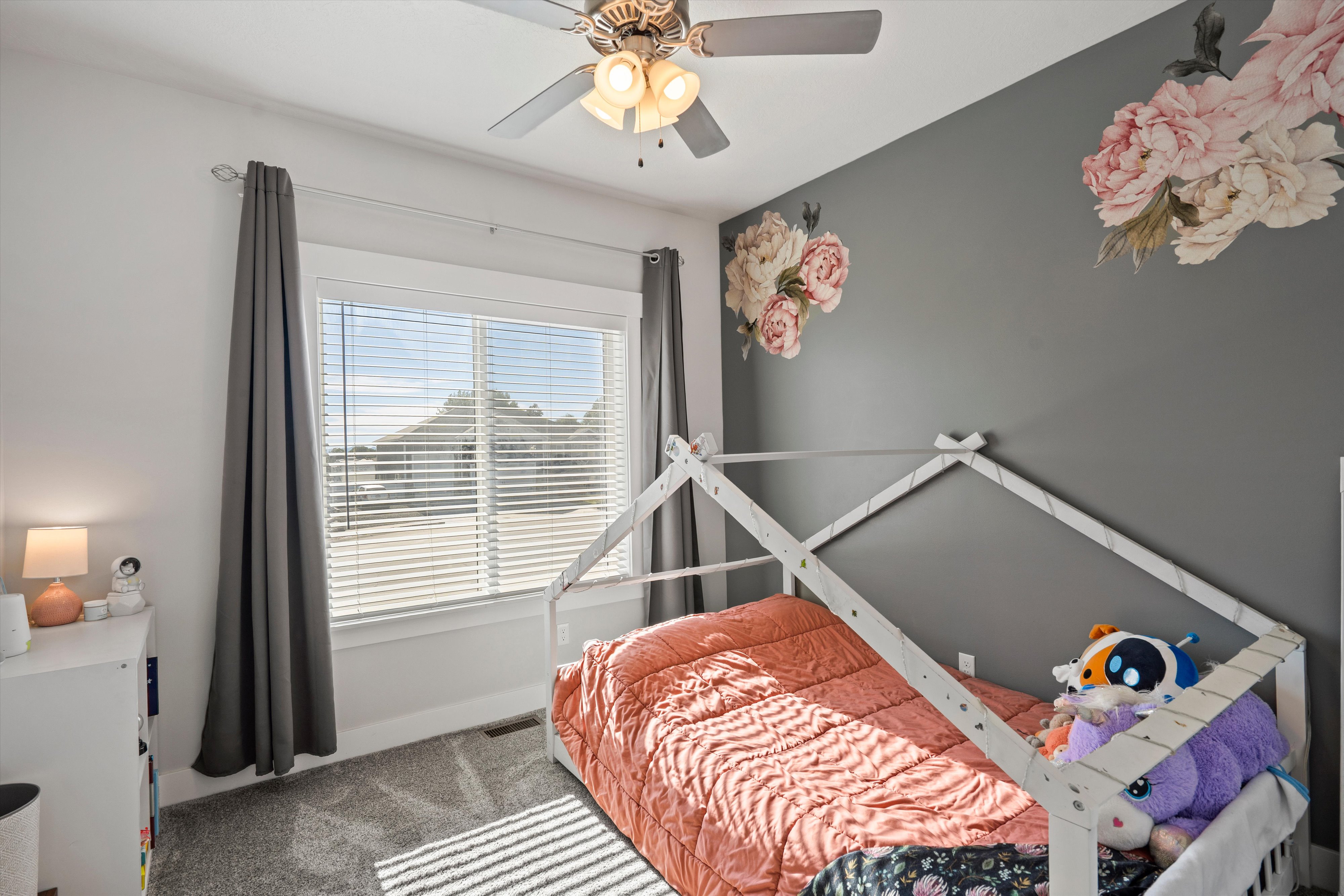 Bedroom featuring a large window with blinds, dark gray wall with floral decals, and a ceiling fan with light fixture