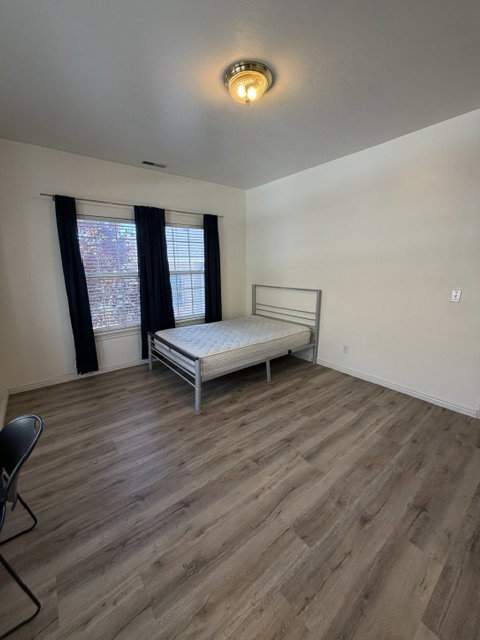Spacious room featuring wood-finish flooring, two windows with blinds, a ceiling-mounted light fixture, and white wall finishes