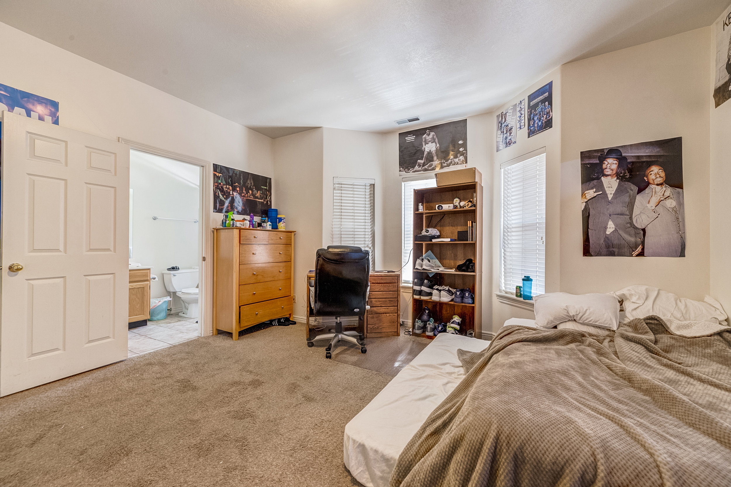 Carpeted bedroom featuring multiple windows with blinds, a doorway to a bathroom with tile flooring and a vanity, and a ceiling light fixture