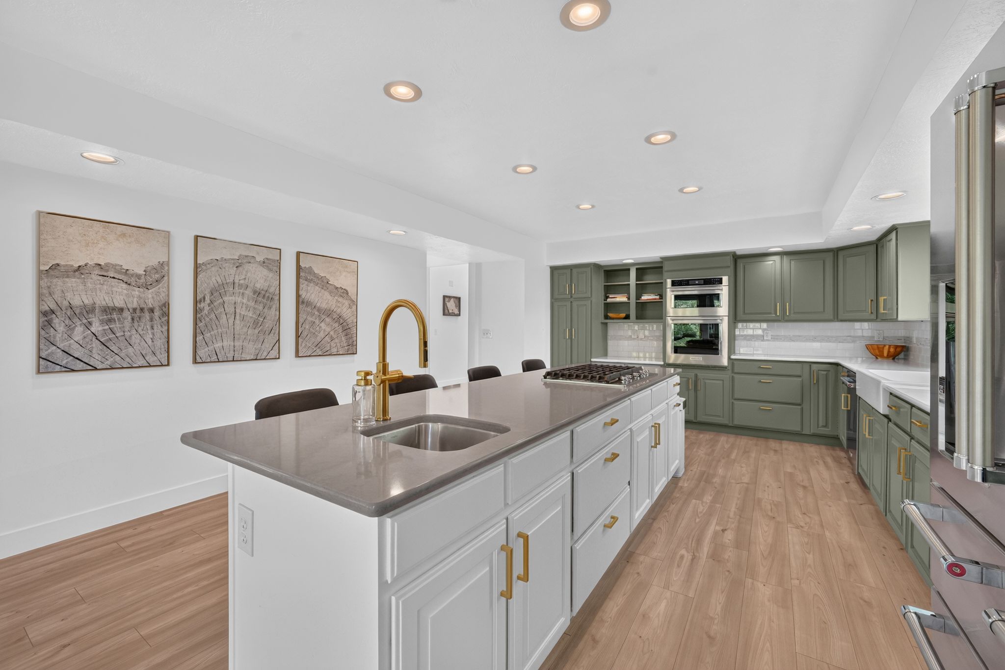 Spacious kitchen featuring a large central island with a gold-tone faucet and integrated sink, light wood-finish flooring, recessed lighting, and sage green cabinetry with gold-tone hardware