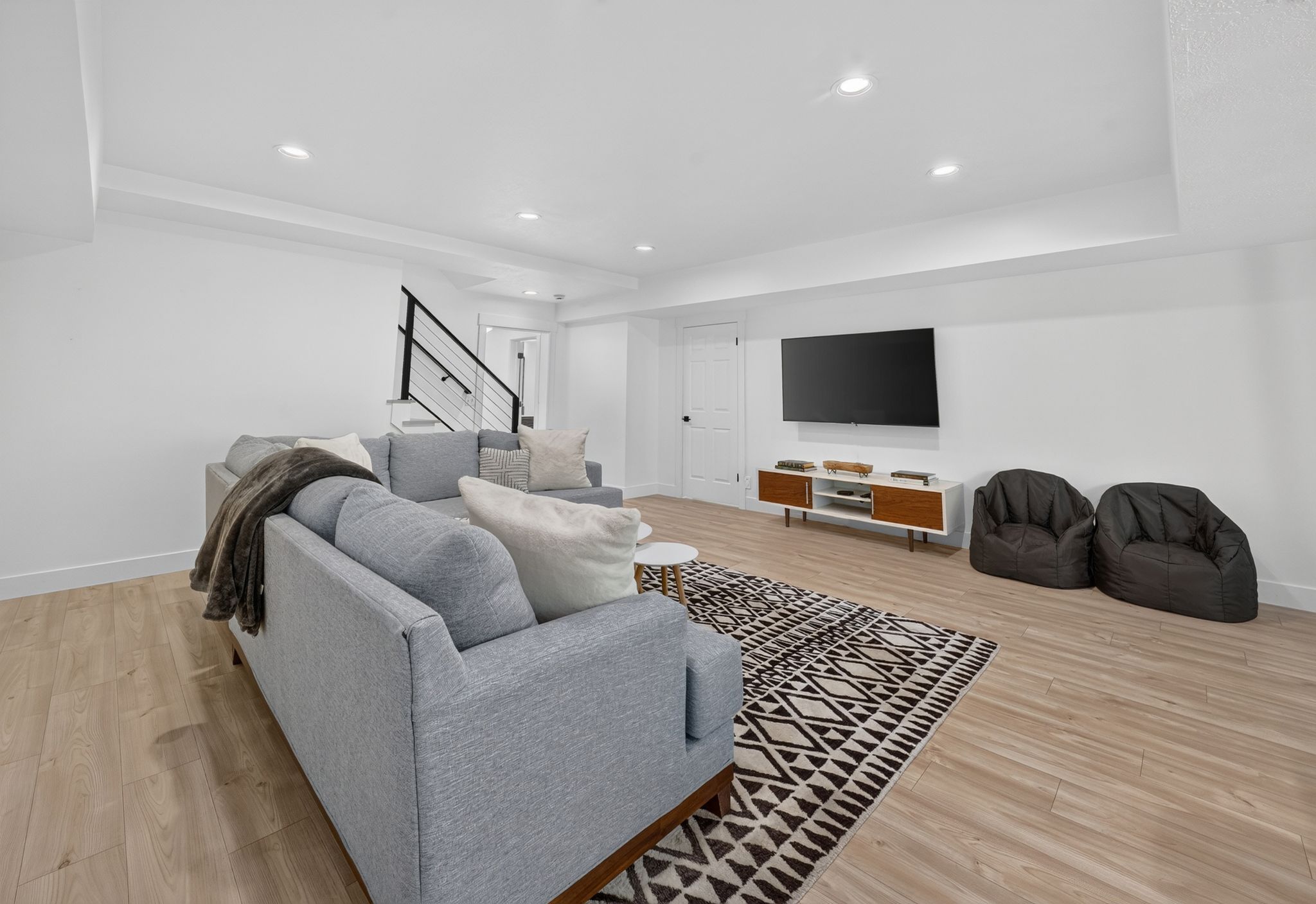 Spacious living area featuring light wood-finish flooring, recessed lighting, and a contemporary black metal staircase railing