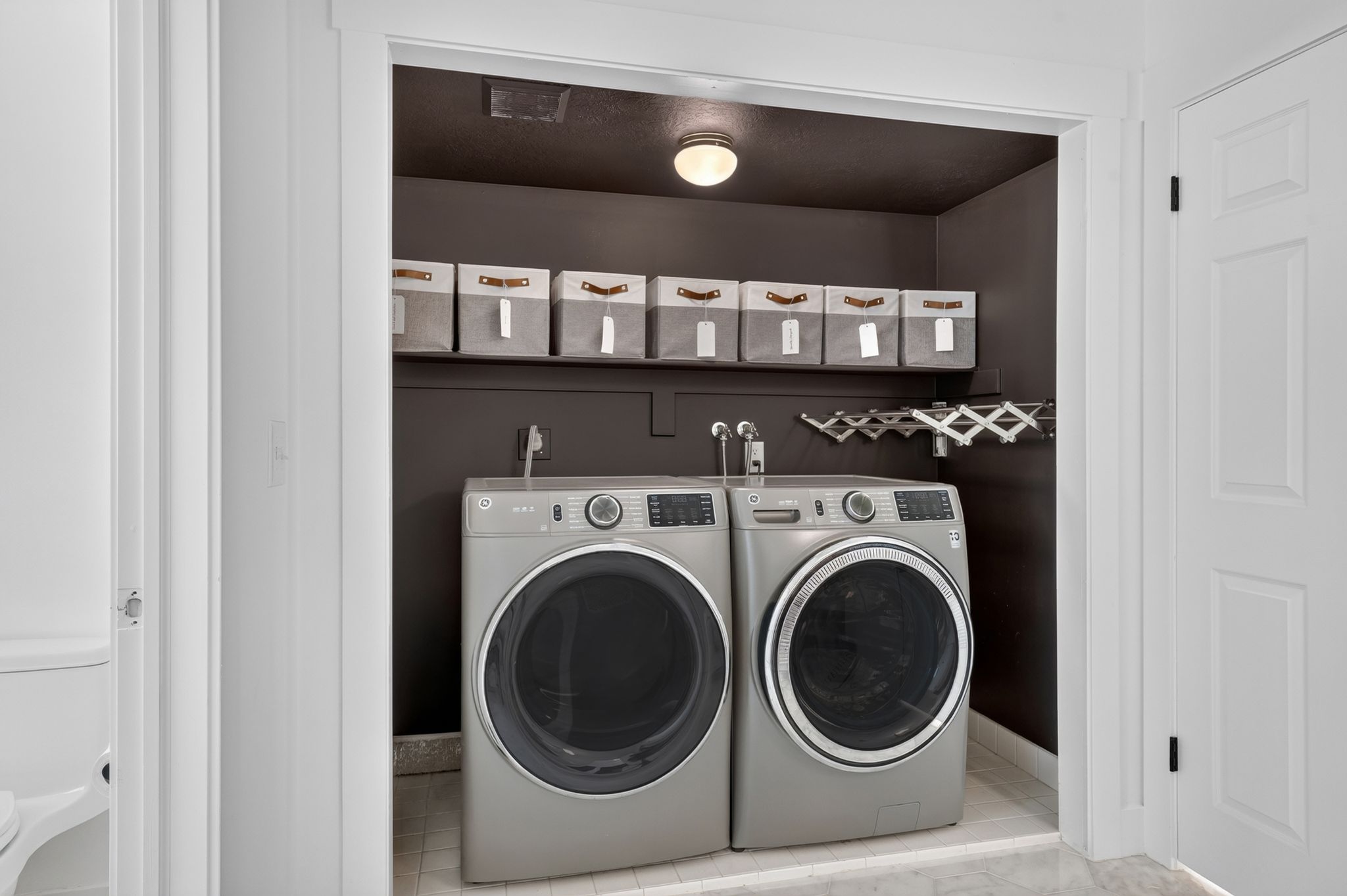 Dedicated laundry area featuring a front-loading washer and dryer, wall-mounted shelving, overhead lighting, and a retractable drying rack
