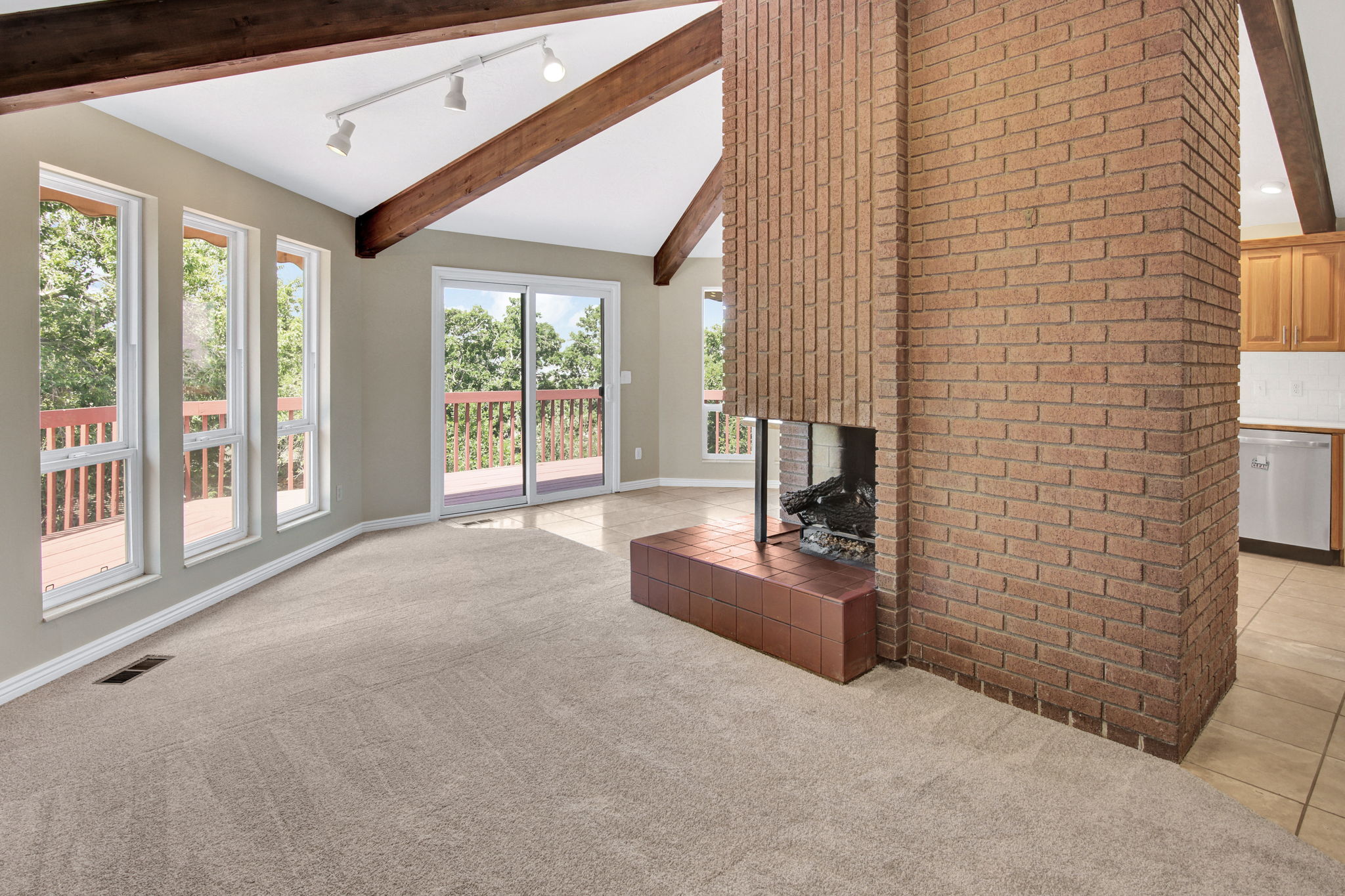 Spacious living area featuring a prominent brick fireplace with a tile hearth, vaulted ceilings with exposed wood beams, and track lighting and view windows. Access huge decking