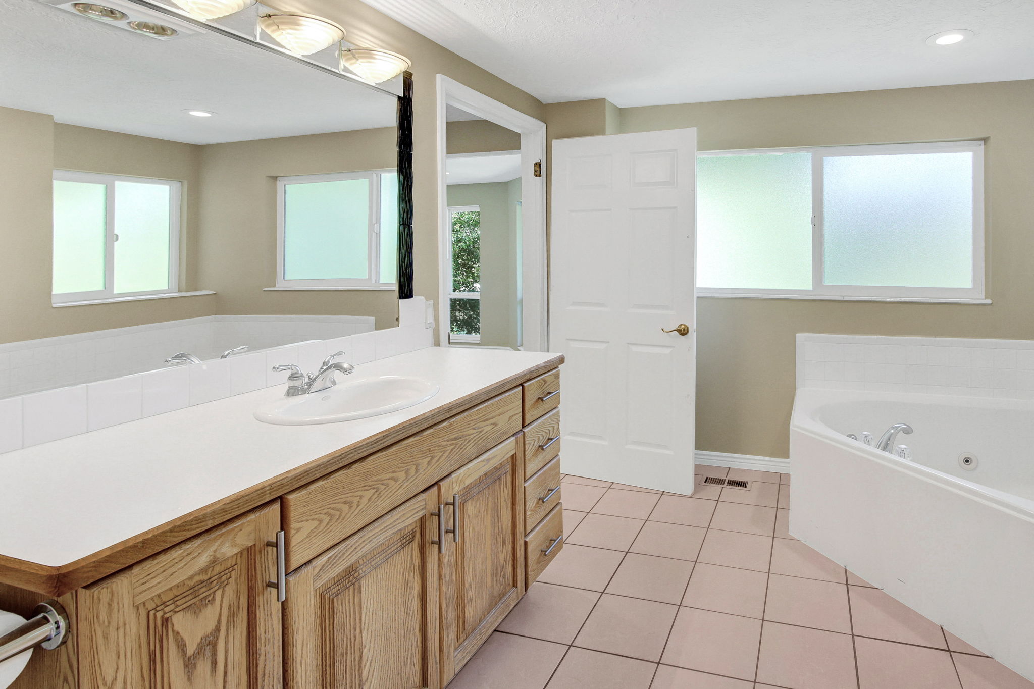 View 2 Ensuite Bathroom featuring a wood-finish vanity with a white countertop and integrated sink, a large wall-mounted mirror, a corner jetted tub, and frosted windows