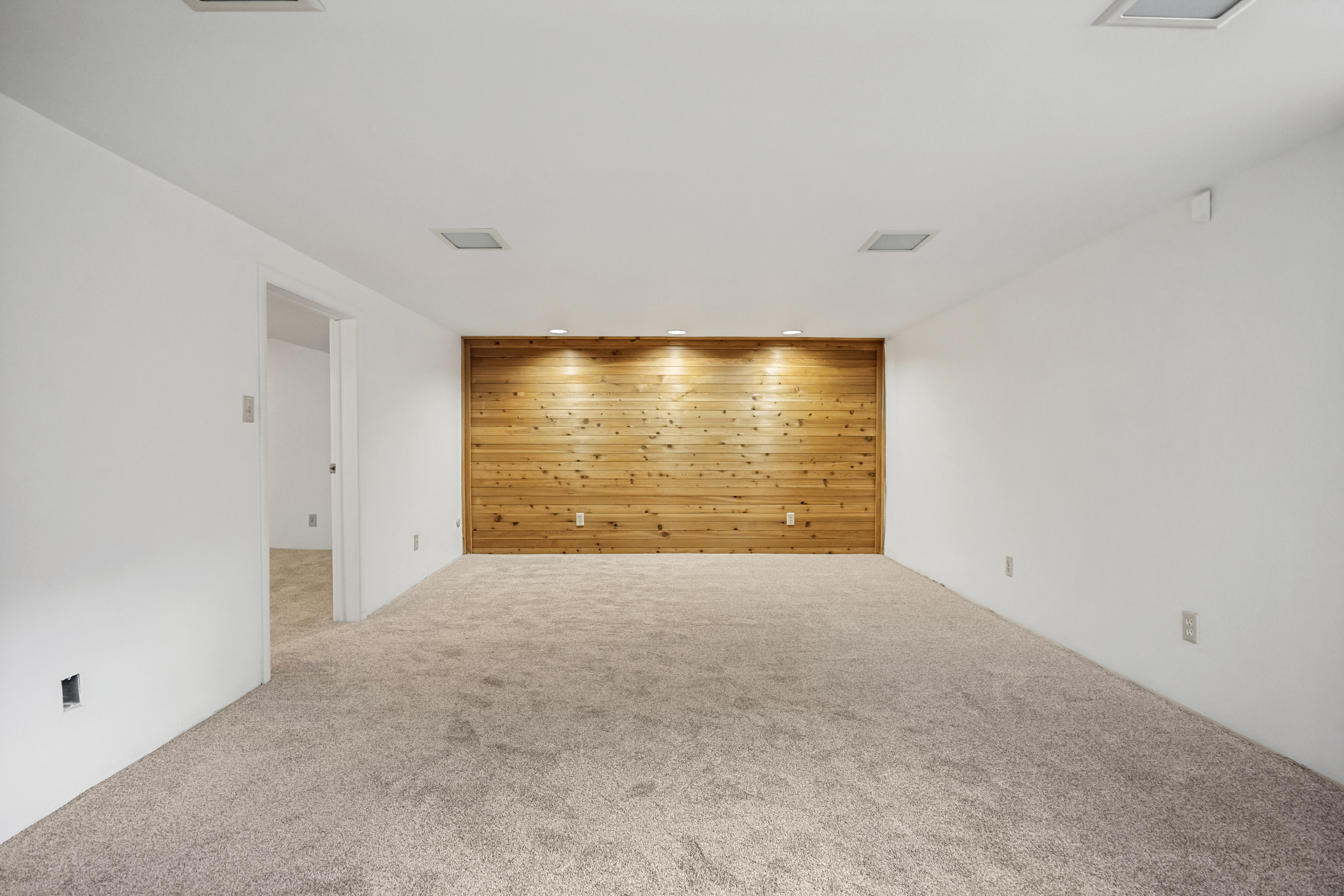 Spacious room featuring a prominent wood accent wall with recessed lighting, light-colored carpeting, white walls, and a doorway opening to an additional room