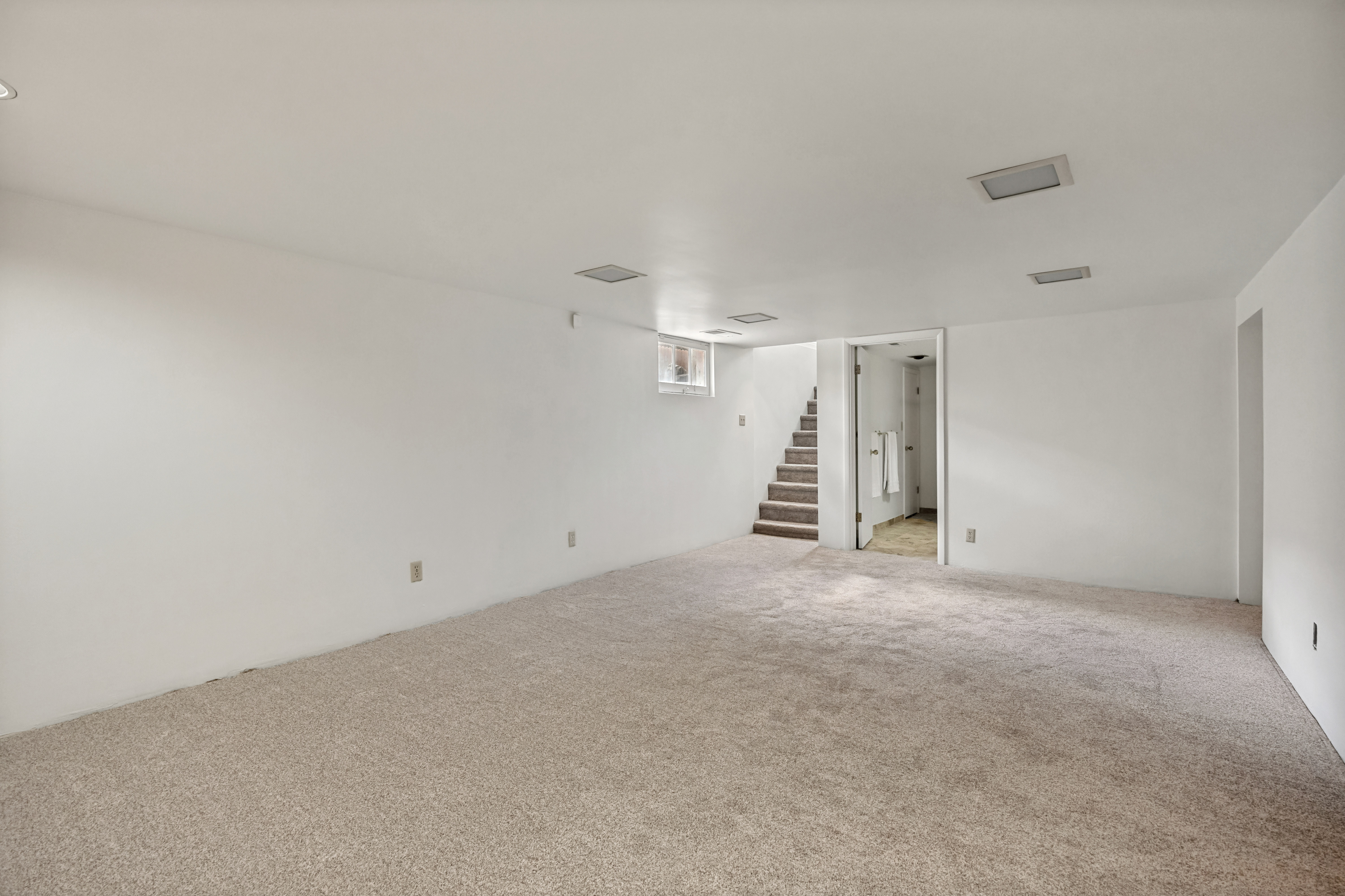 Expansive carpeted area with neutral-toned walls, recessed lighting, and a visible staircase providing access to an upper level