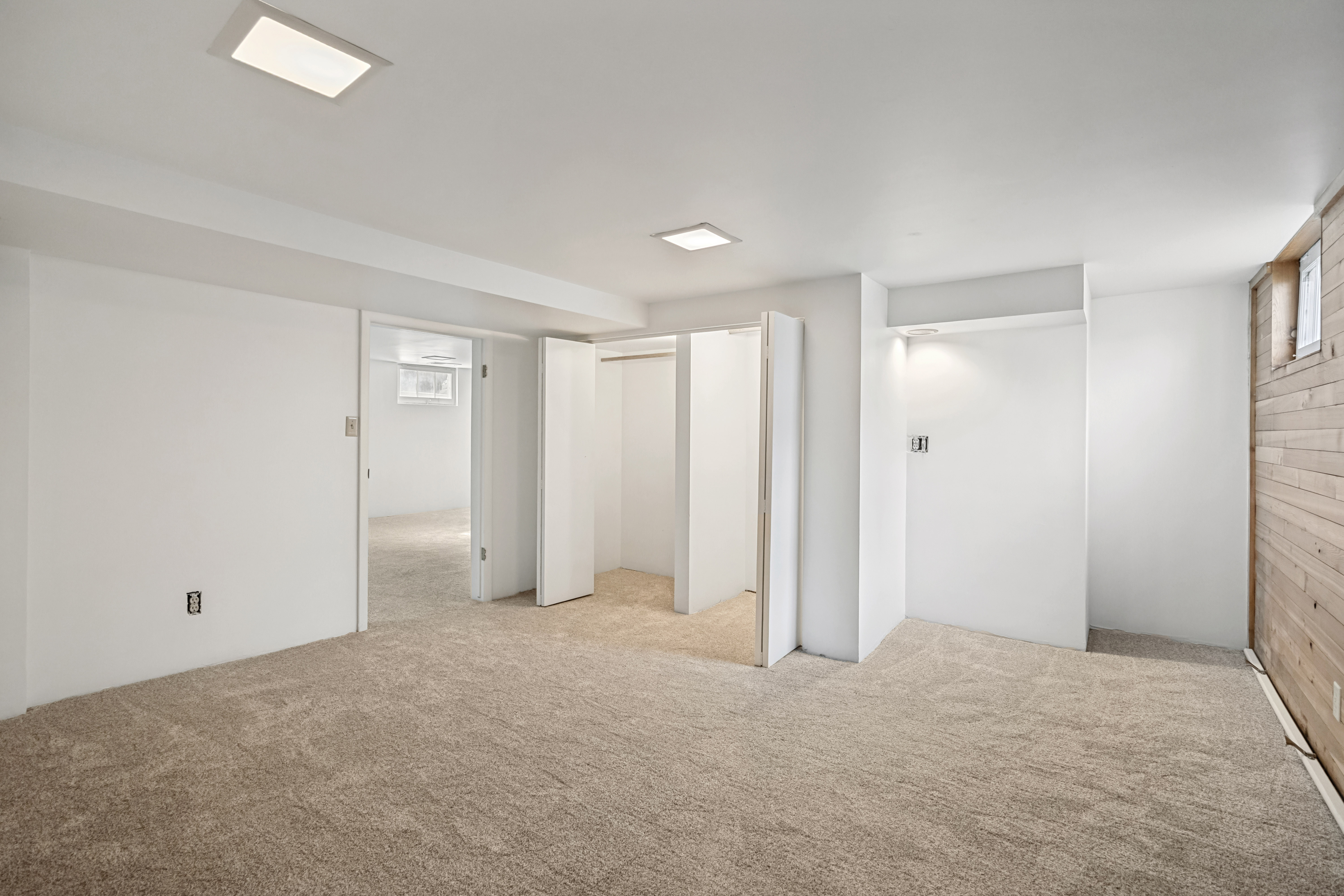 Recessed lighting illuminates a carpeted room featuring a wood-paneled accent wall, multiple closet spaces with bi-fold doors, and a doorway opening to an additional carpeted area