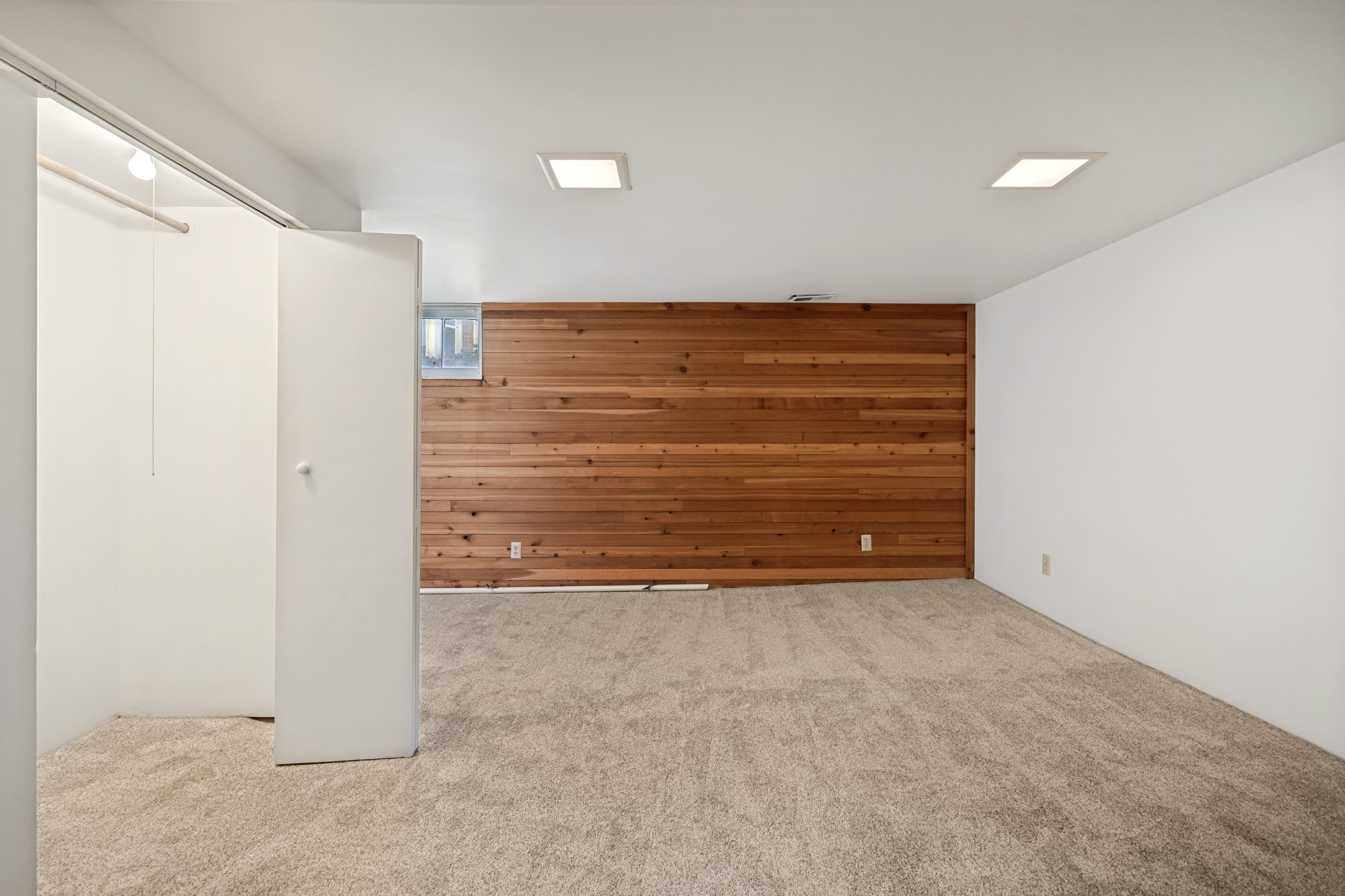 Spacious room featuring a wood-paneled accent wall, neutral carpeting, flush-mount lighting, and an integrated closet with bi-fold doors