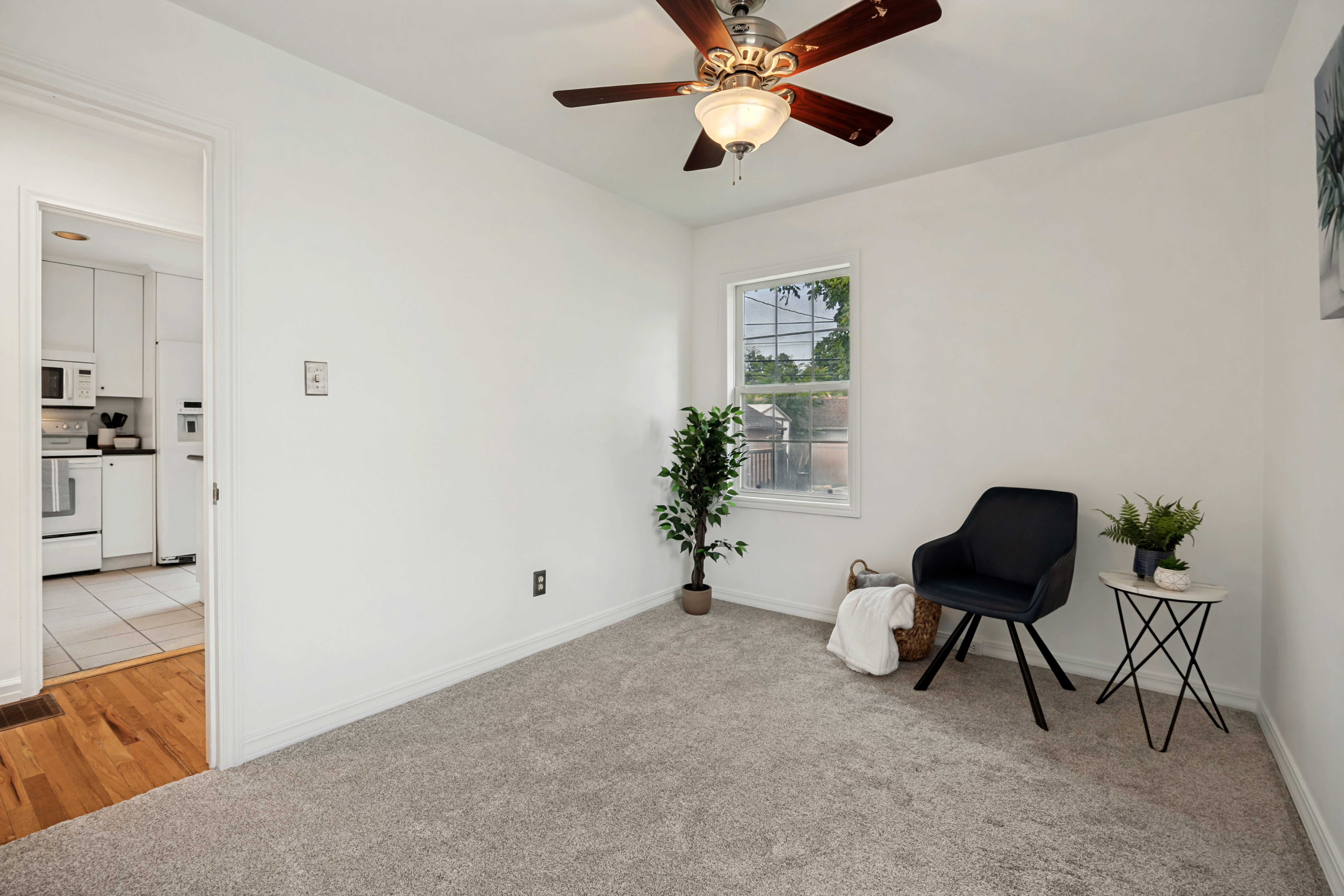 Carpeted room with a ceiling fan, a window, and an open doorway revealing a kitchen with white cabinetry, white appliances, and tiled flooring