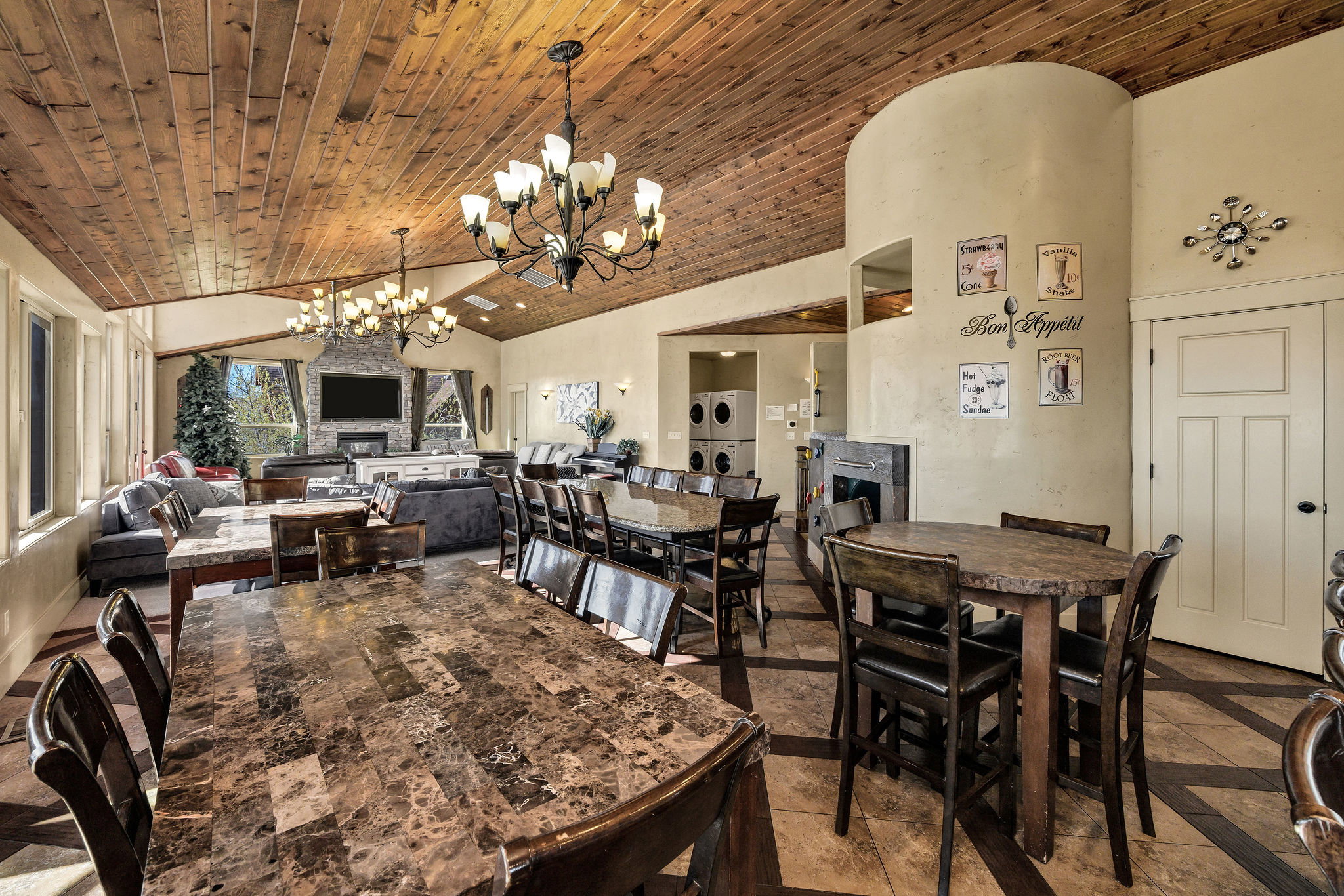 Vaulted wood-plank ceiling with multiple chandeliers, tile flooring, stone-front fireplace, large windows, and a built-in appliance stack