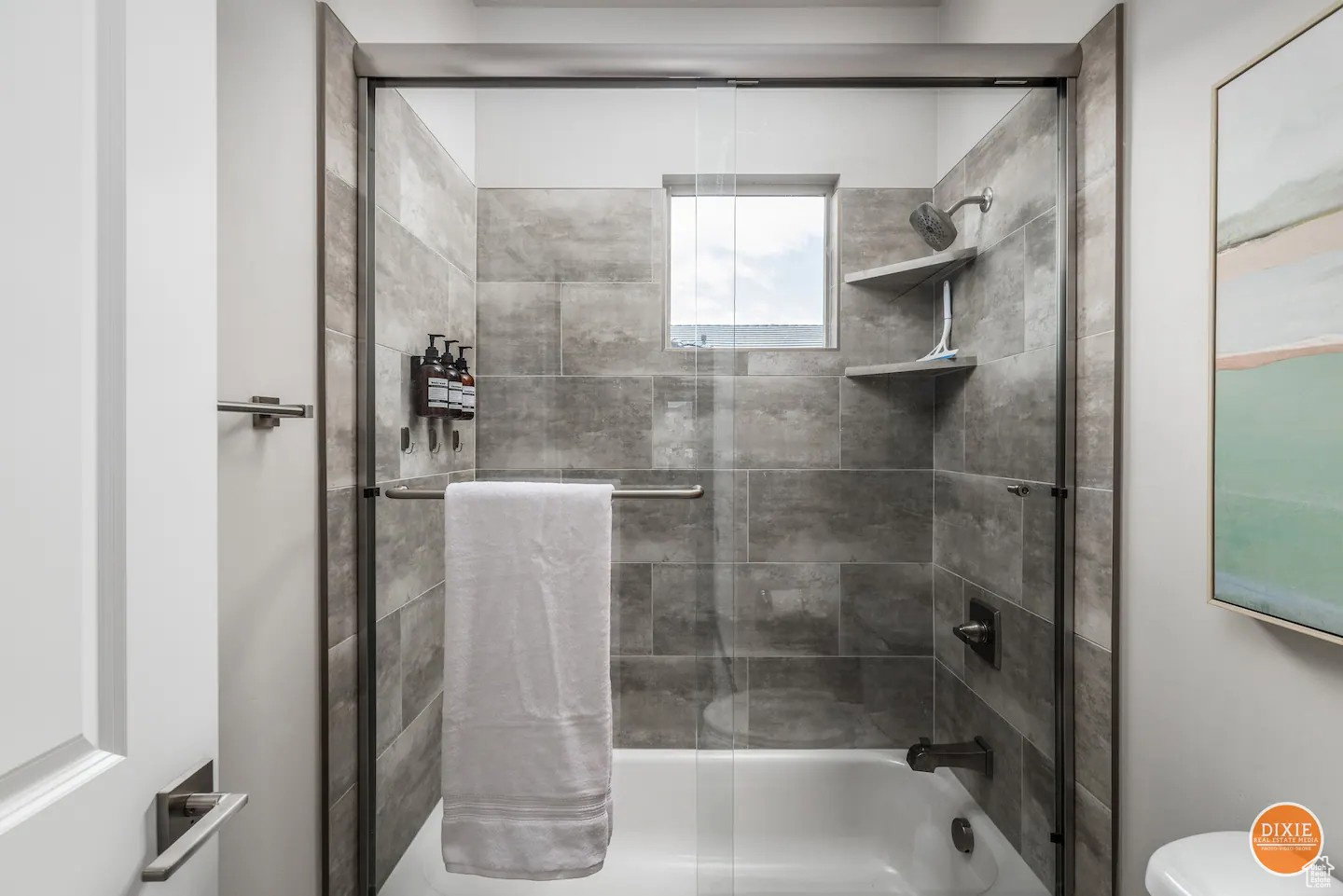 Contemporary tub and shower combination featuring a glass enclosure with a sliding door, textured gray tile surround, built-in shelving, and a window providing natural light