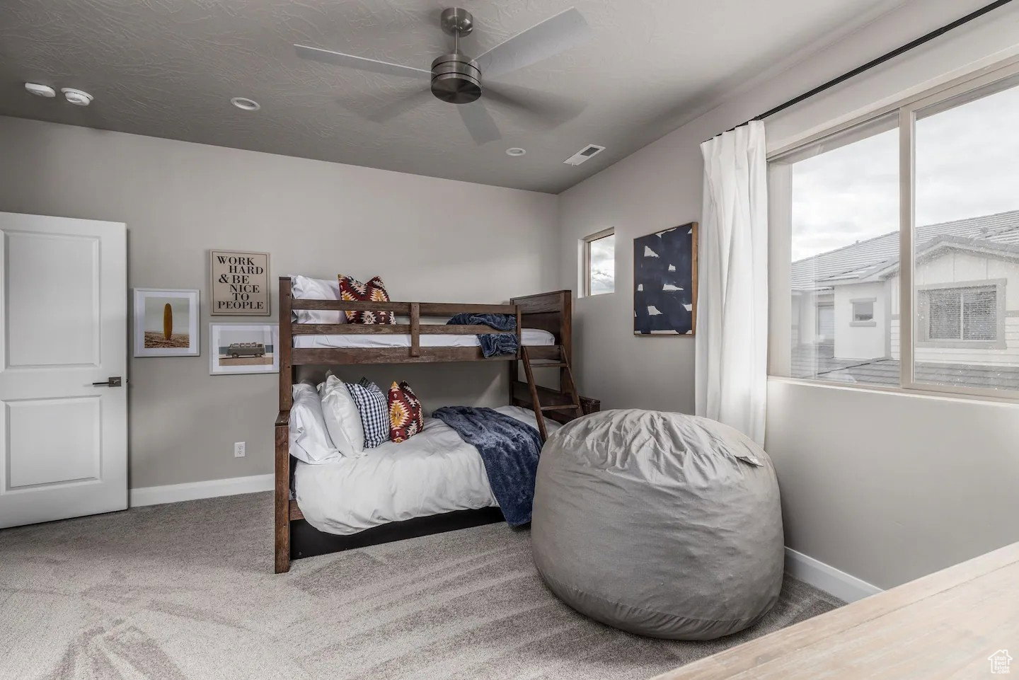Spacious room featuring neutral-toned walls, patterned carpet flooring, a ceiling fan with integrated lighting, and multiple windows allowing natural light
