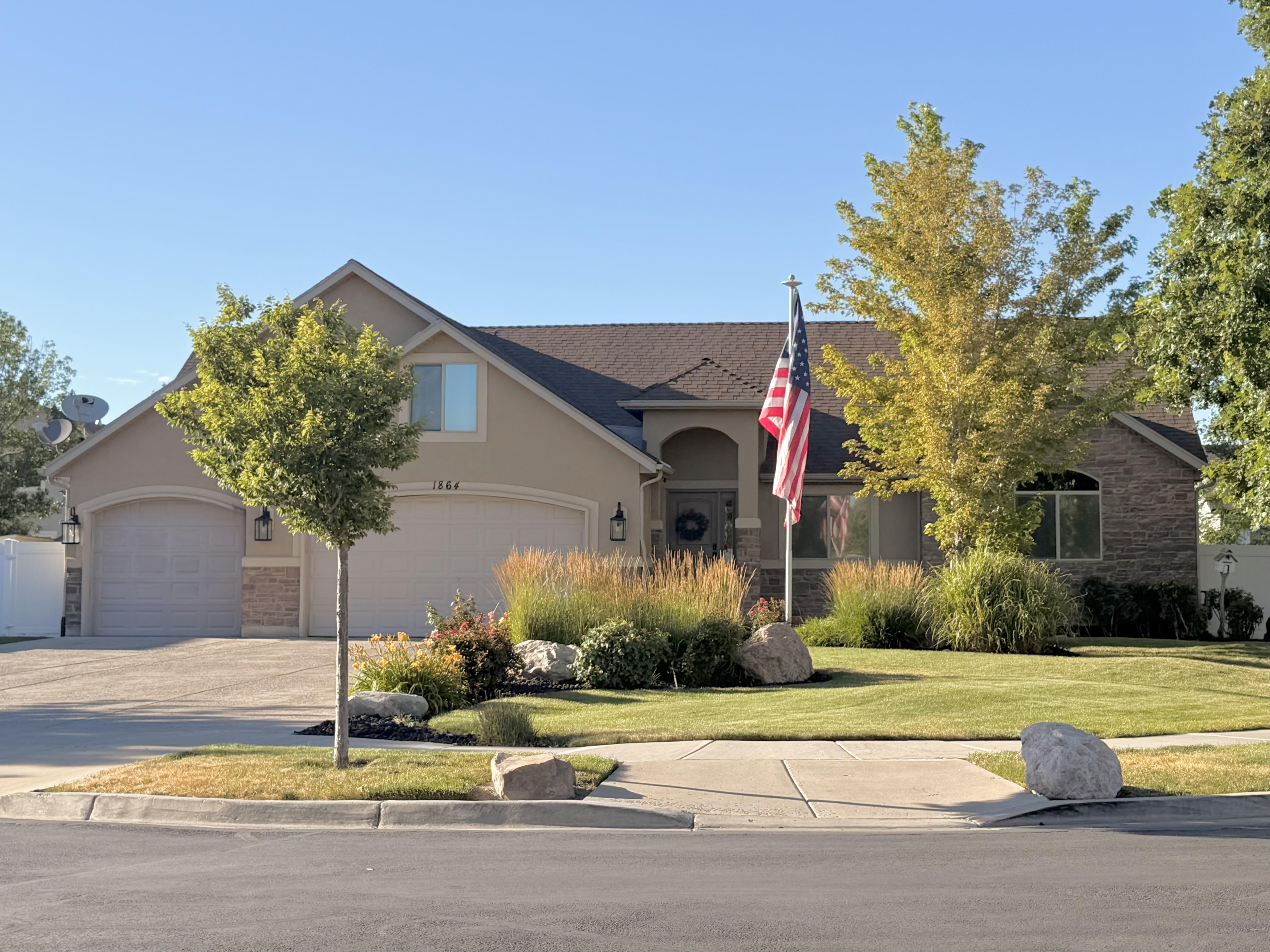 Exterior featuring a two-car garage, stucco and brick facade, front lawn, mature trees, and a decorative landscape bed with rocks
