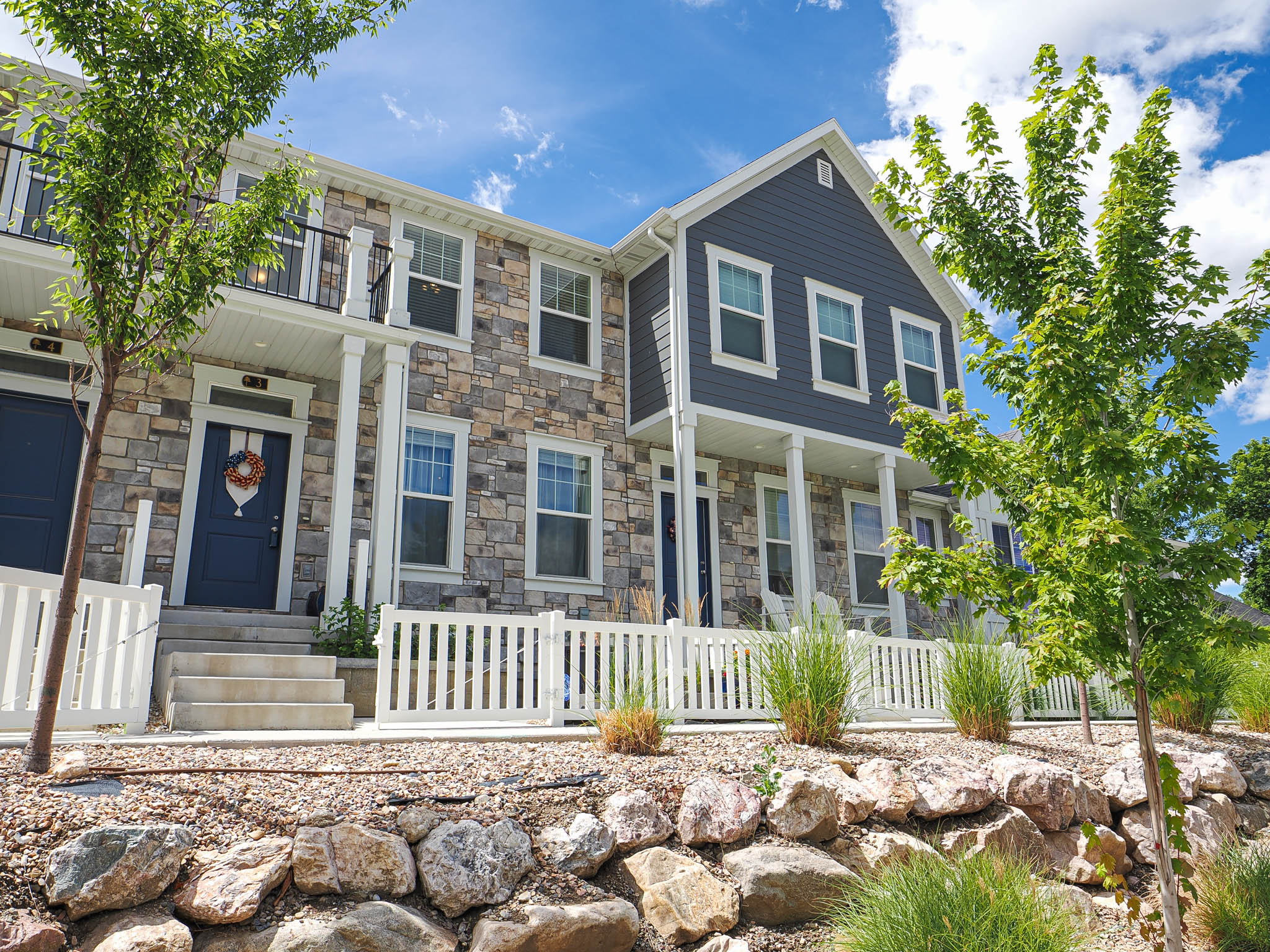 Townhome exterior featuring stone and siding finishes, a white picket fence, a covered porch, an upper-level balcony with black railing, and mature landscaping with rockery