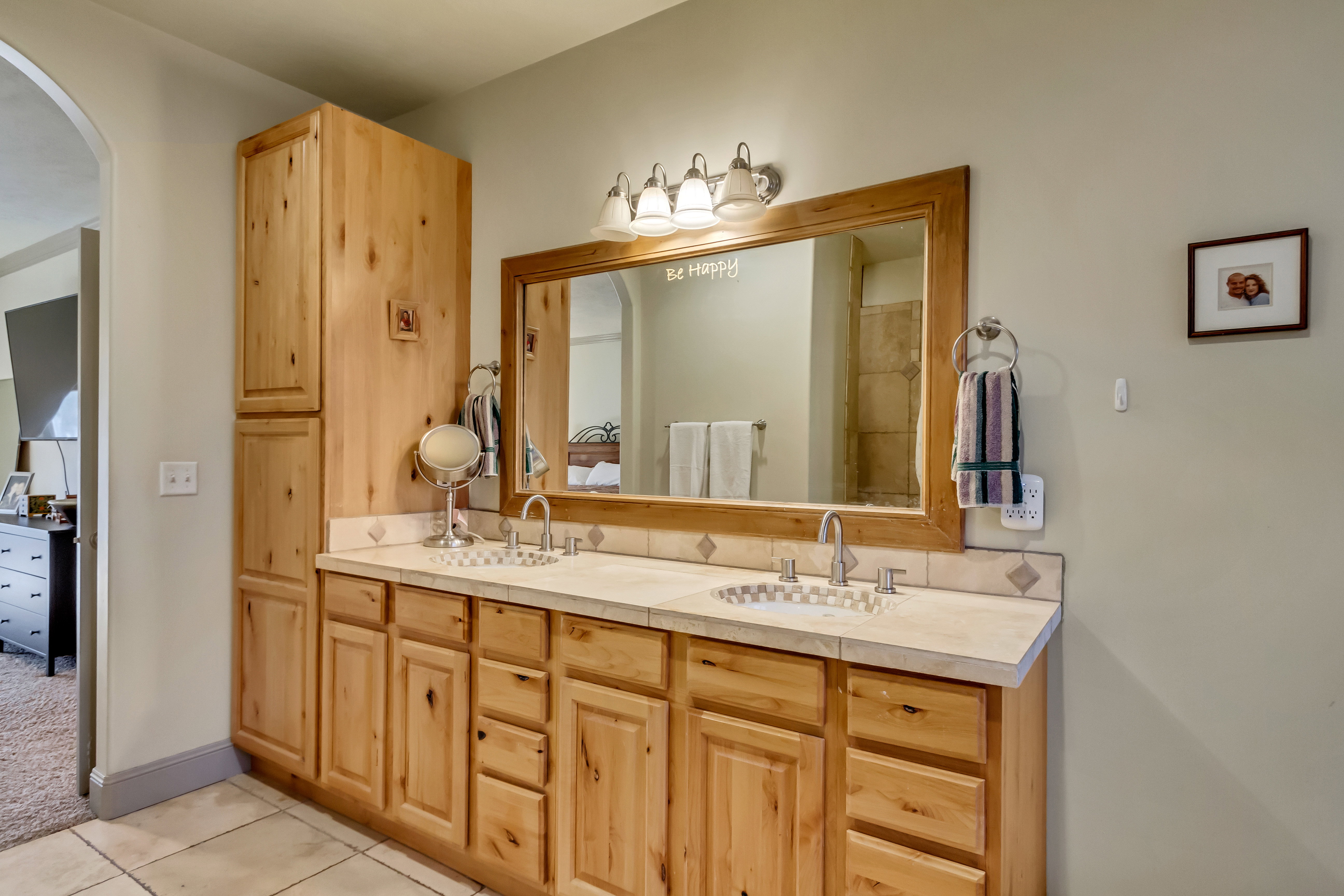 Dual sink vanity with light wood-finish cabinetry, a framed mirror, and a tile countertop with integrated sinks