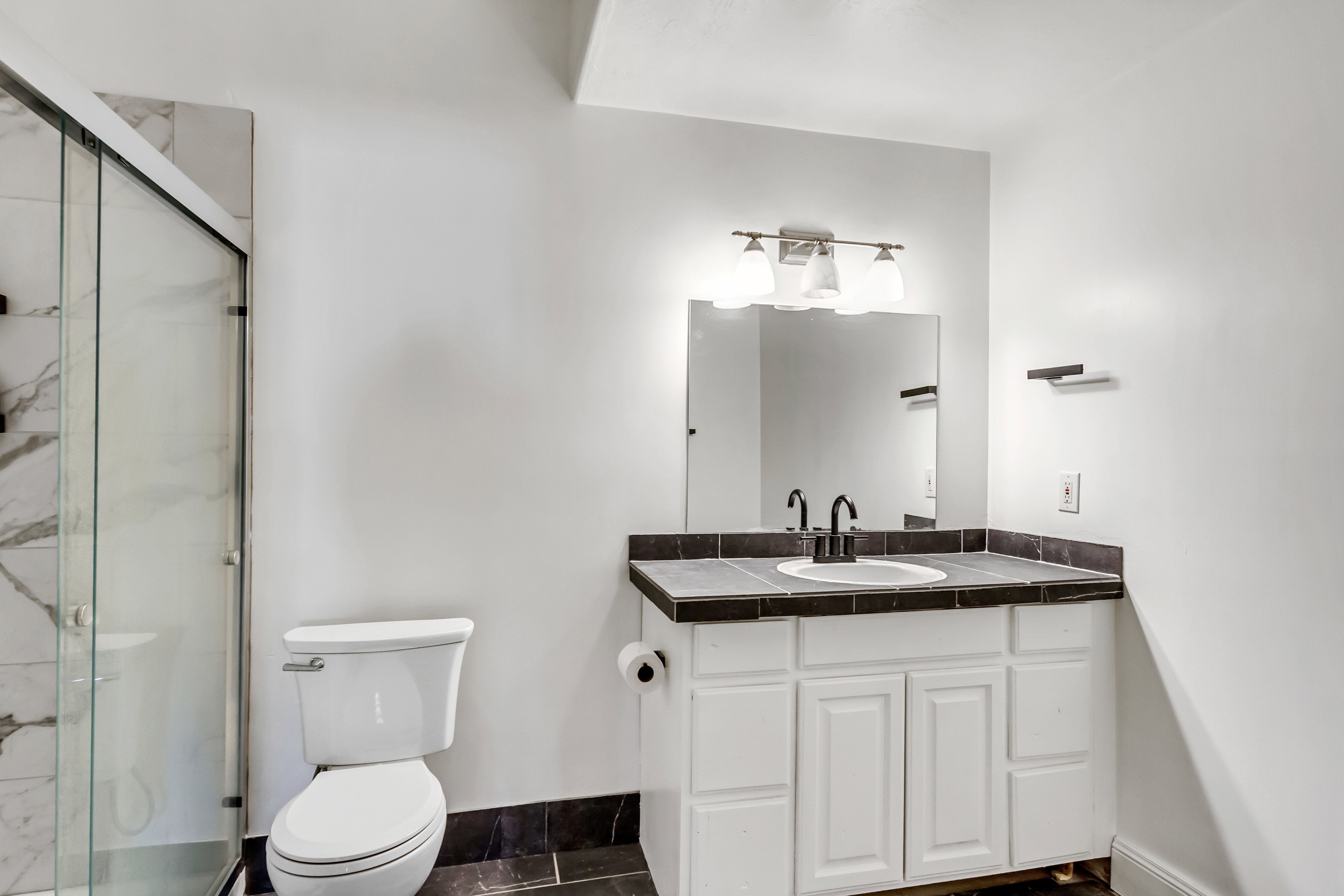 Bathroom featuring a glass-enclosed shower with marble-look tile, white vanity with a dark tiled countertop and integrated sink, black fixtures, and a wall-mounted mirror with overhead lighting