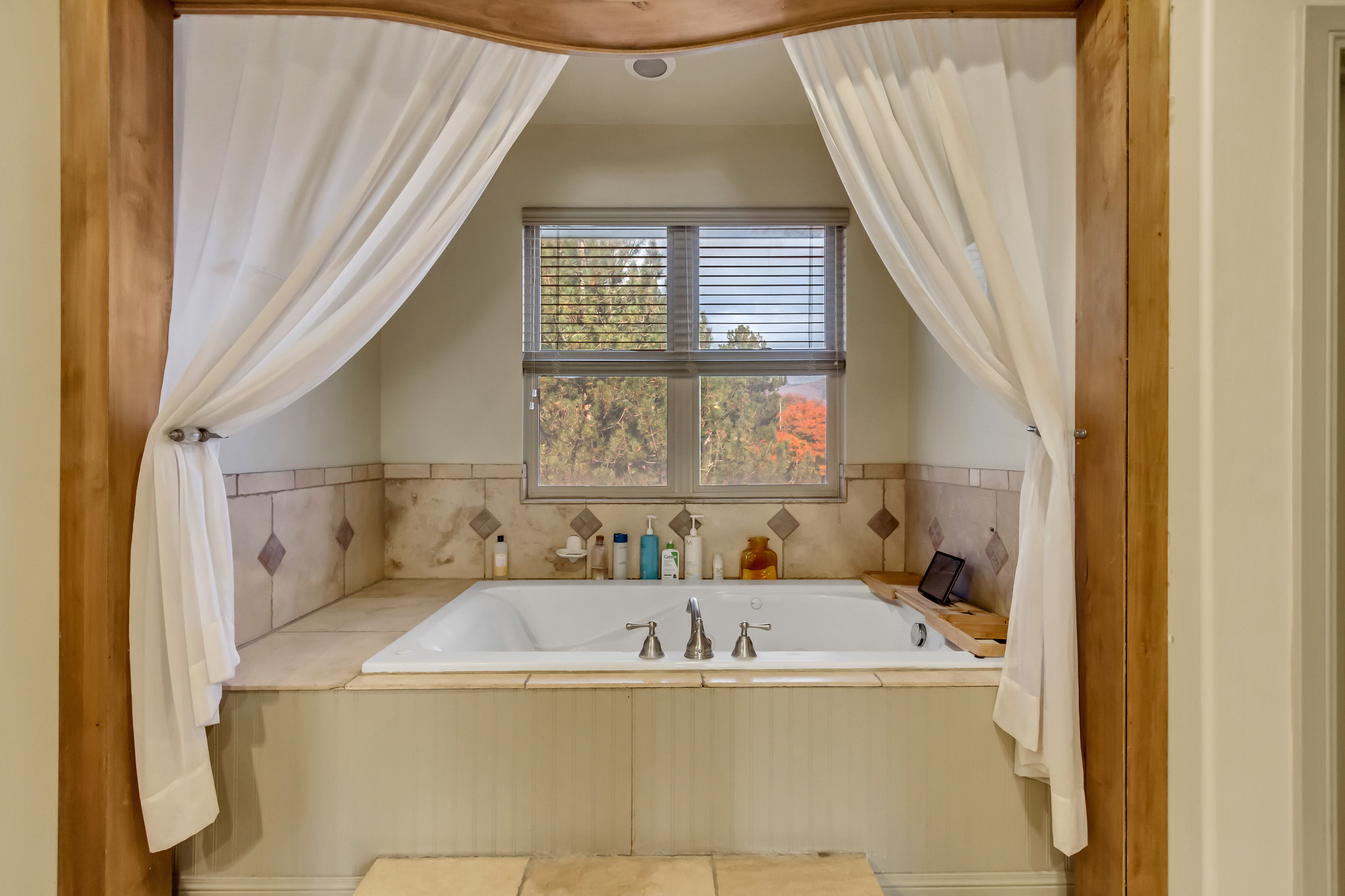 Built-in soaking tub with deck-mounted faucet, surrounded by tiled walls with diamond accents, and a window with horizontal blinds
