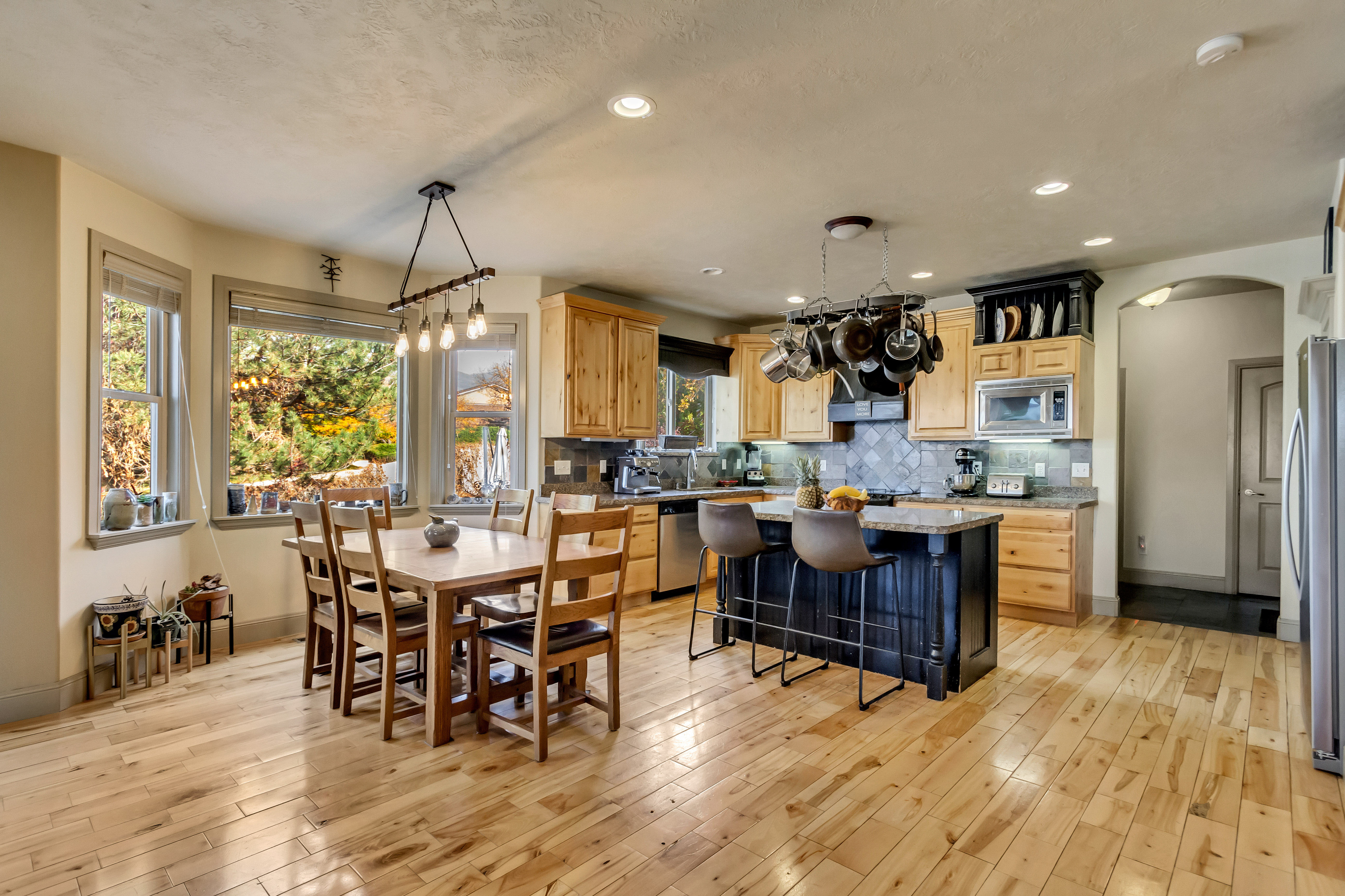 Open-concept kitchen and dining area featuring light wood-finish flooring, a central island with a dark base and seating, light wood cabinetry, stainless steel appliances, and multiple windows