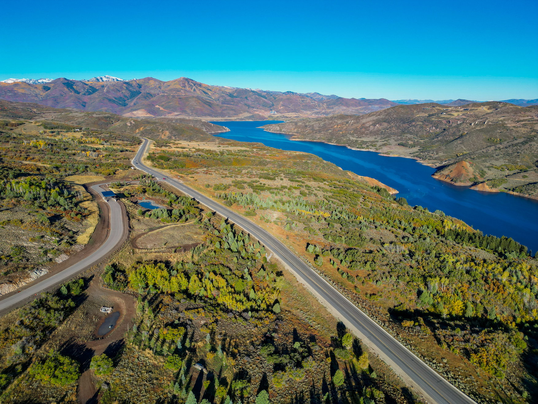 Expansive mountain and water scenery featuring a paved road, a serene body of water, and distant snow-capped peaks