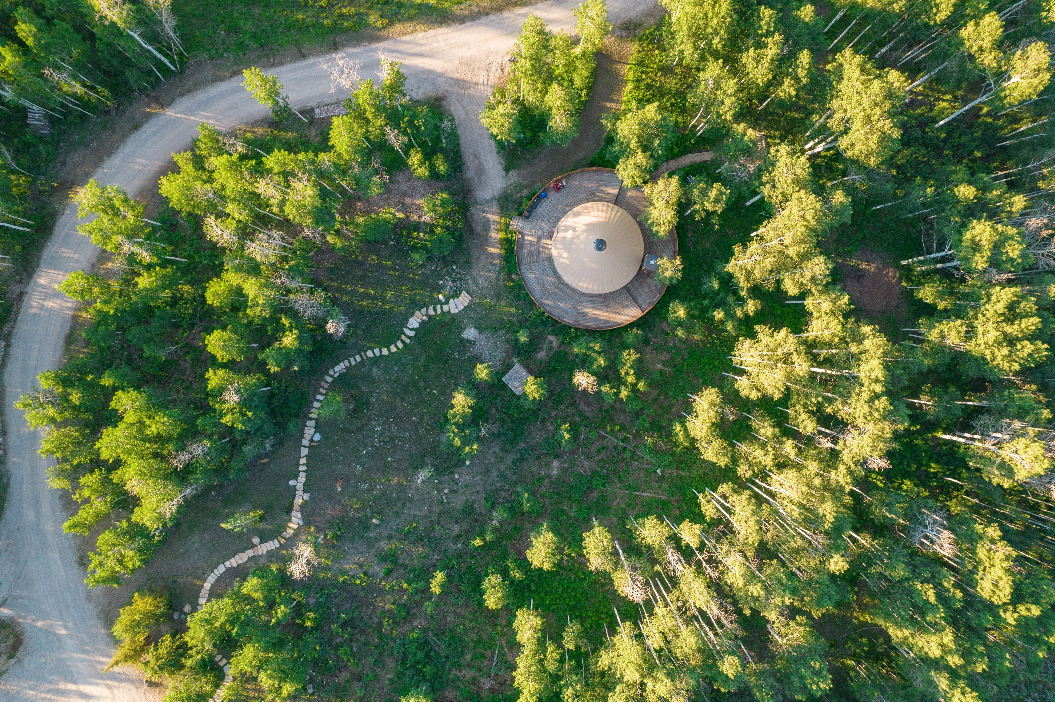 Circular dwelling surrounded by dense deciduous and evergreen trees, featuring a winding stone pathway, a circular wooden deck, and a gravel drive