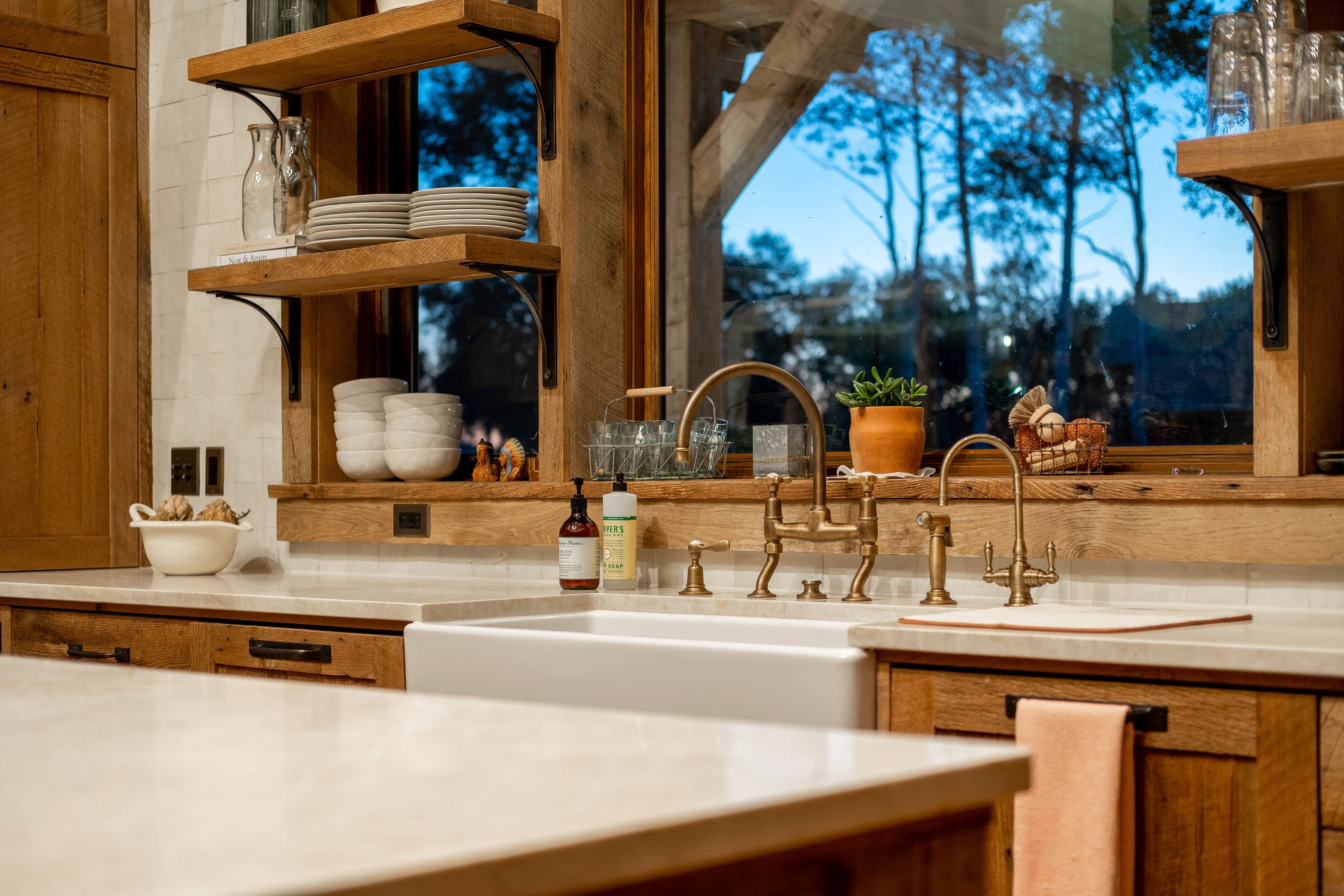 Kitchen featuring a white farmhouse sink with brass fixtures, wood-finish cabinetry, light-toned countertops, and open shelving