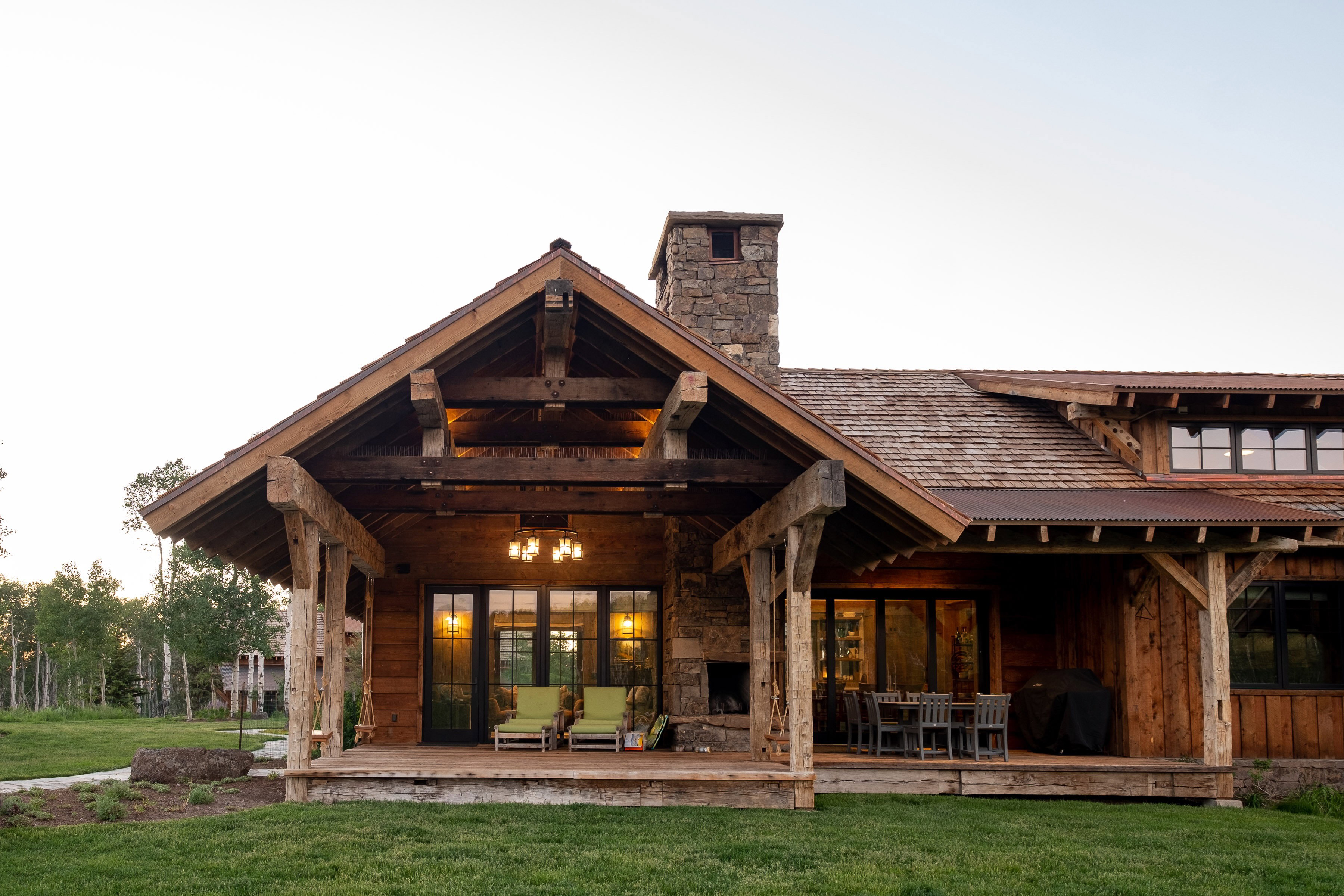 Expansive covered patio featuring a stone fireplace, exposed timber beams, wood-finish siding, a shake shingle roof, and multiple large windows