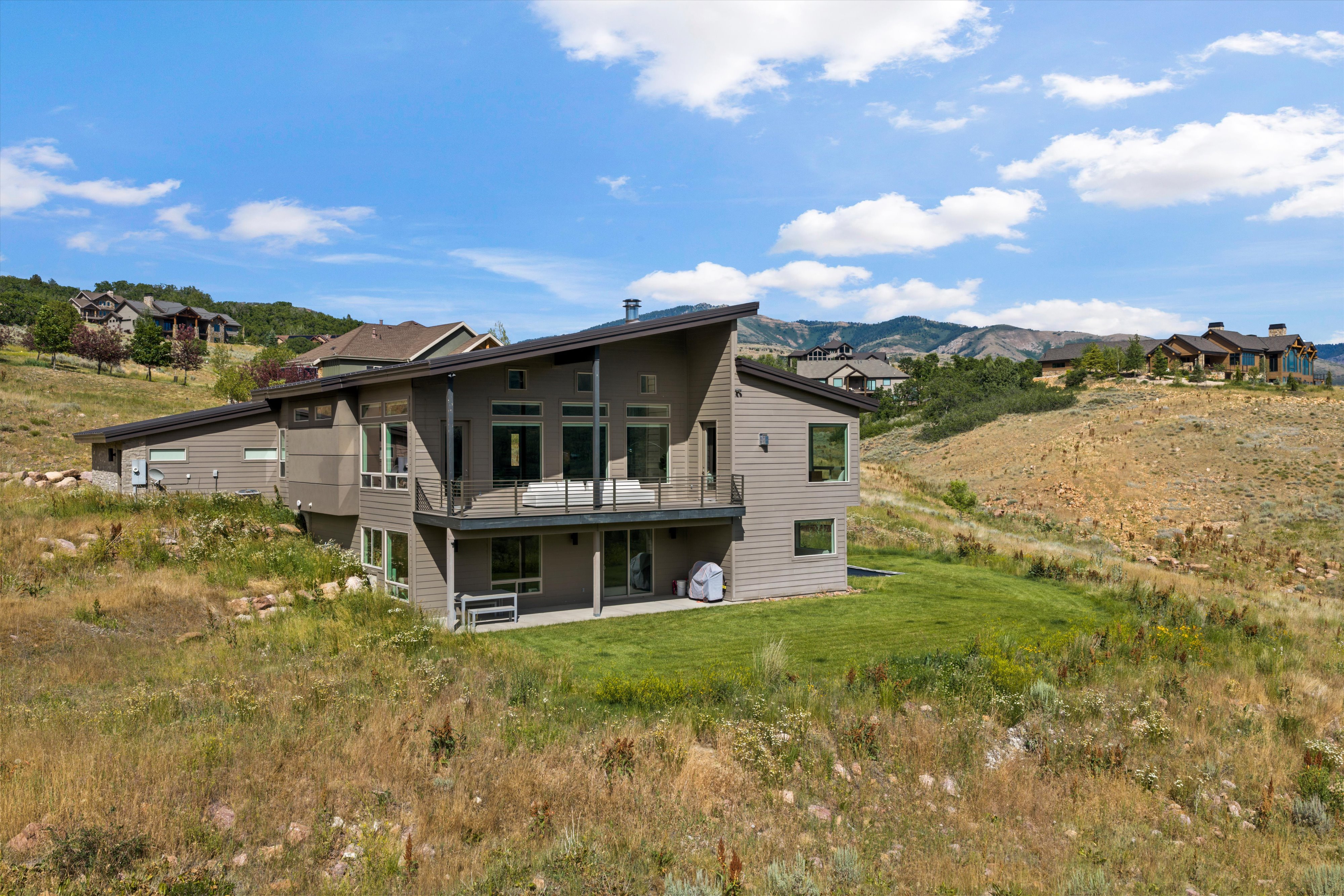 Contemporary multi-level residence featuring a sloped roofline, a second-story balcony with glass railings, an outdoor patio, expansive windows, and a neutral-toned siding exterior