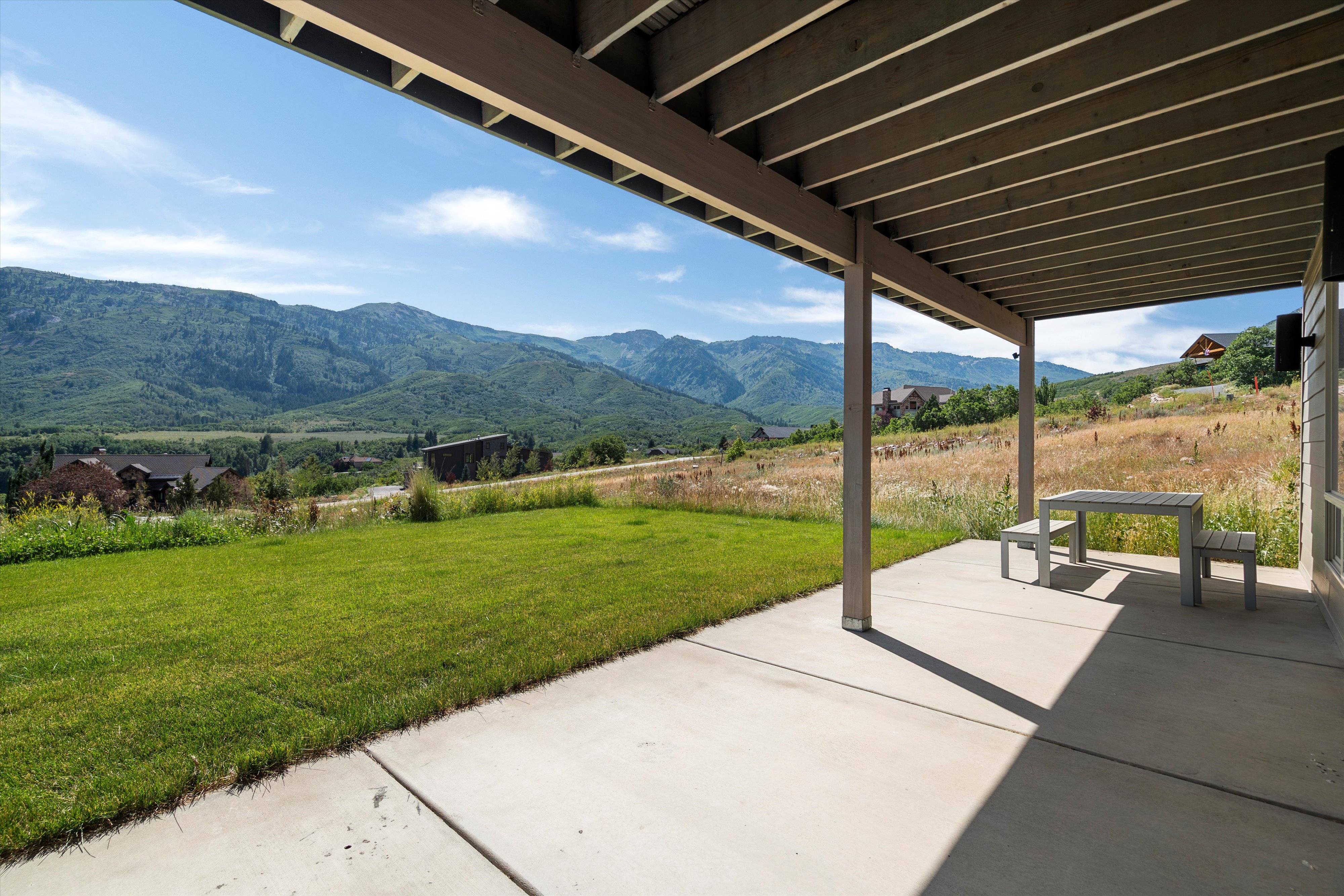 Covered outdoor patio with concrete flooring and exposed beam ceiling, opening onto a grassy lawn