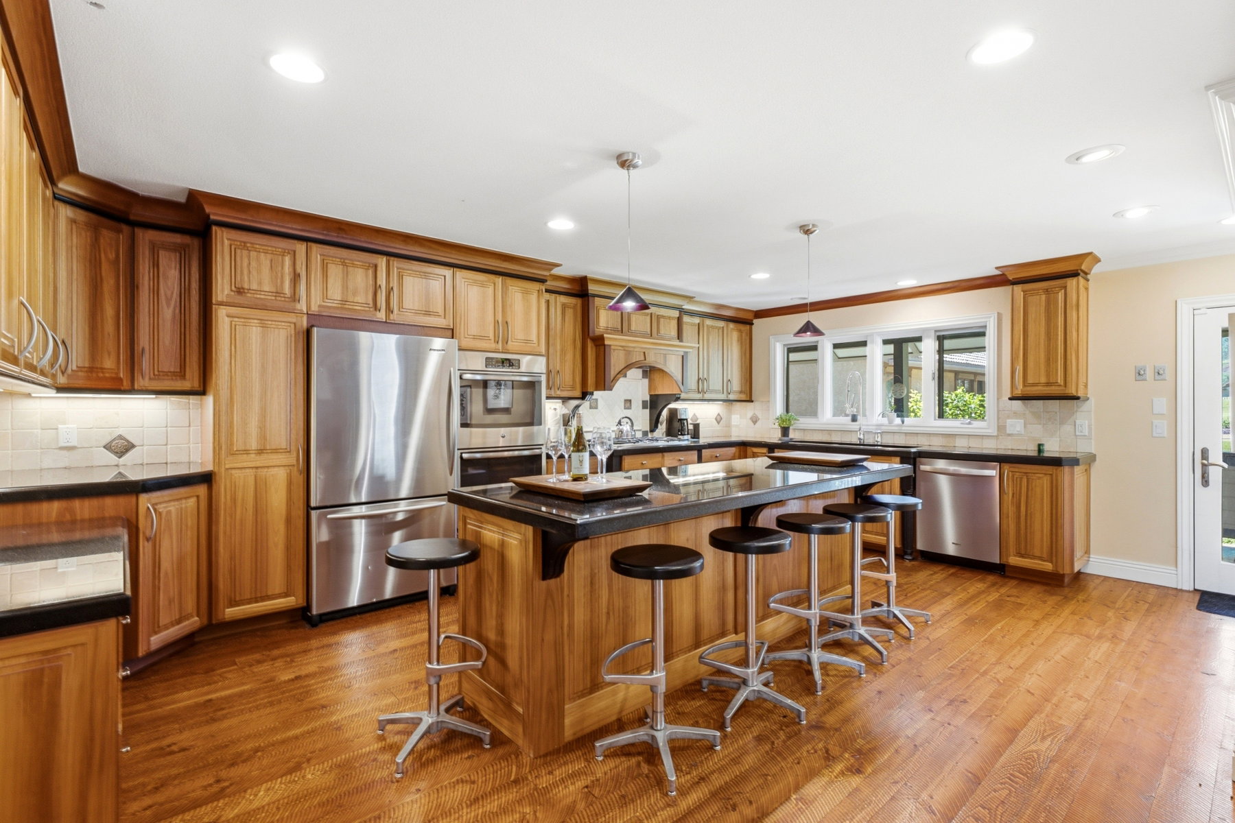 Kitchen featuring a central island with seating, wood-finish flooring, custom wood cabinetry, stainless steel appliances, and a tile backsplash