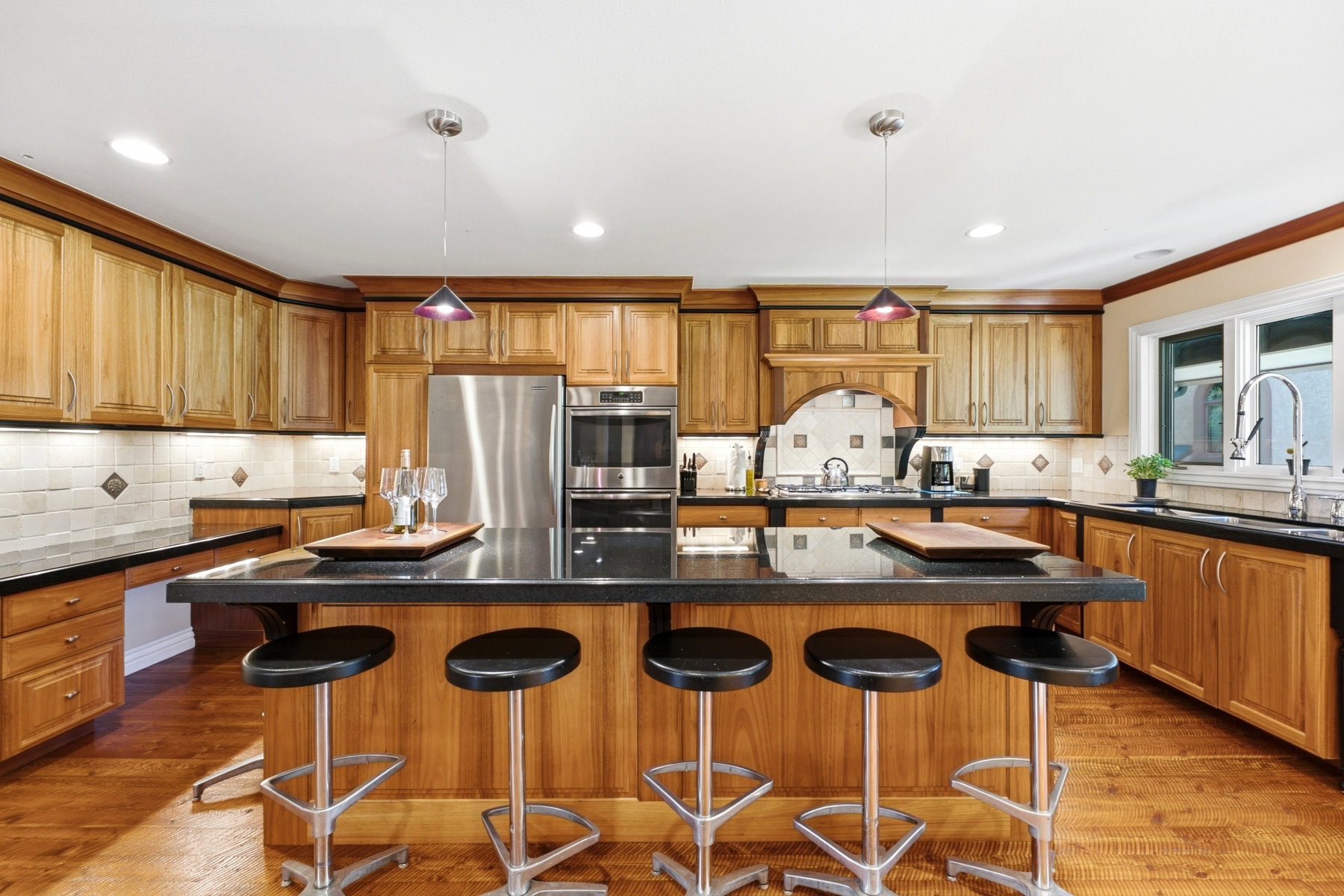 Expansive kitchen featuring a large central island with seating, natural wood cabinetry, dark countertops, tile backsplash with decorative accents, and stainless steel appliances