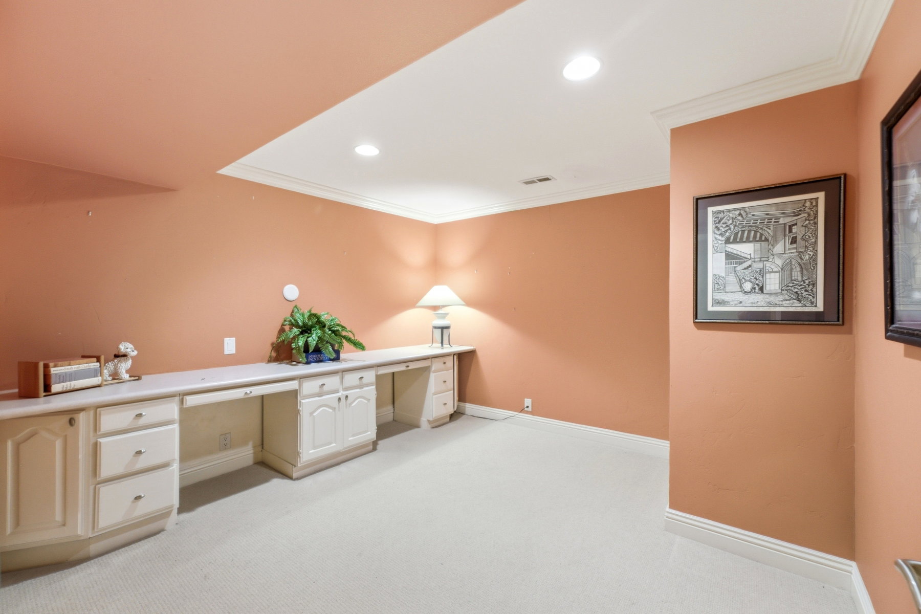 Expansive room featuring a lengthy built-in desk with multiple drawers and cabinets, recessed ceiling lighting, crown molding, and plush carpeting