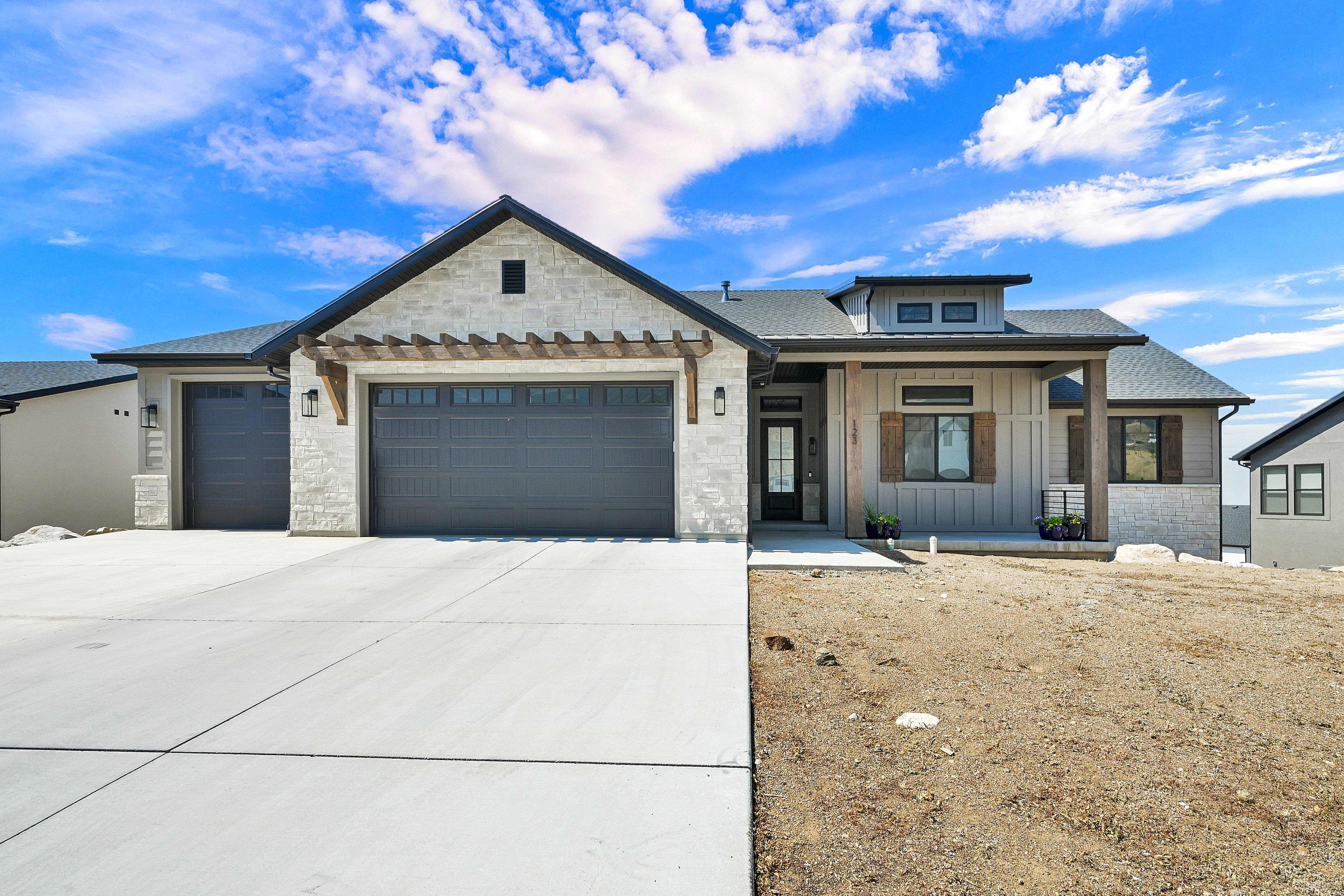 Contemporary ranch-style exterior featuring stone and vertical siding accents, a prominent front porch with wood-finish columns, a two-car garage with pergola detailing, and an additional single-car garage