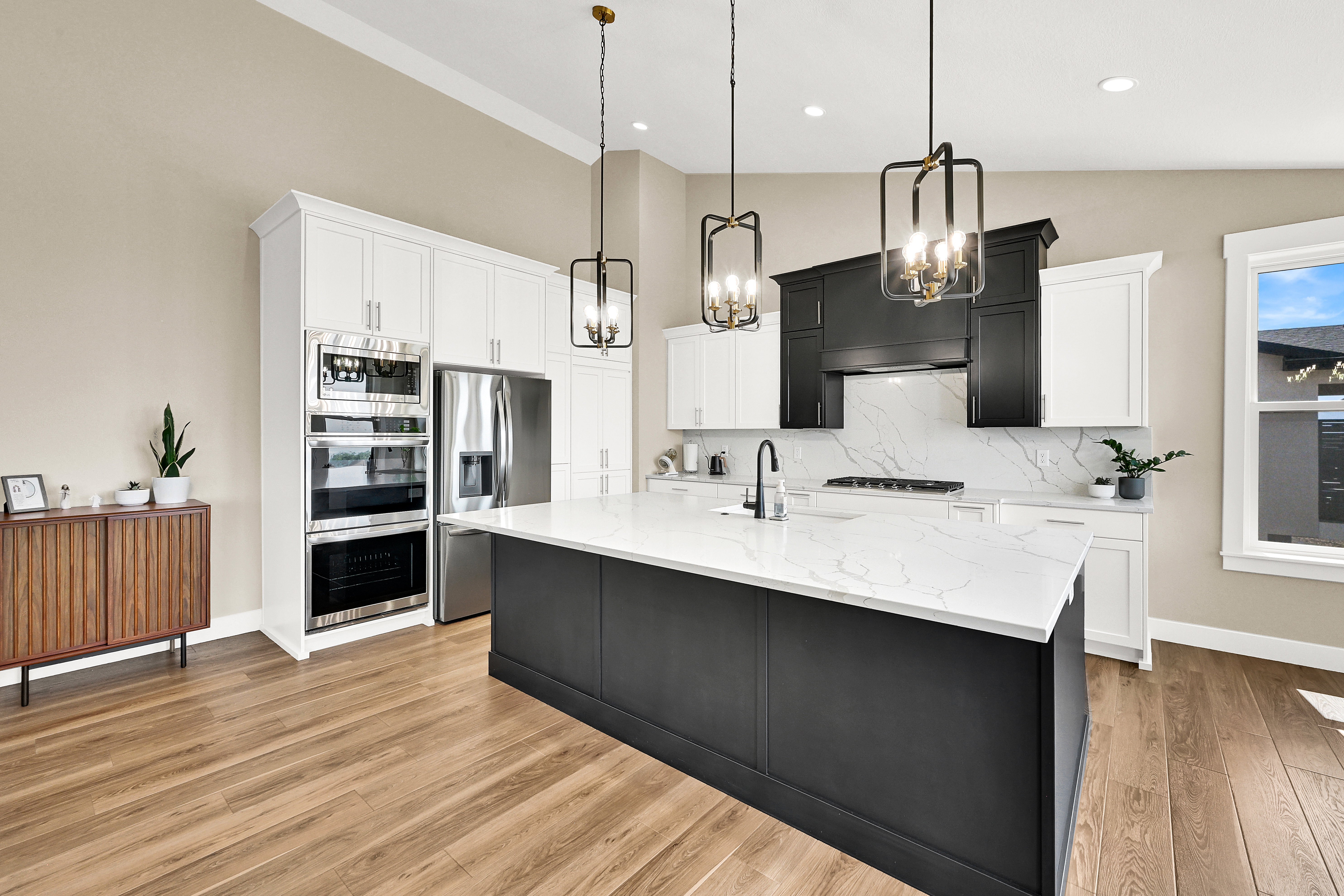 Spacious kitchen featuring a large island with a light-toned countertop, stainless steel appliances, wood-finish flooring, white perimeter cabinetry, and black accent cabinetry with a matching hood