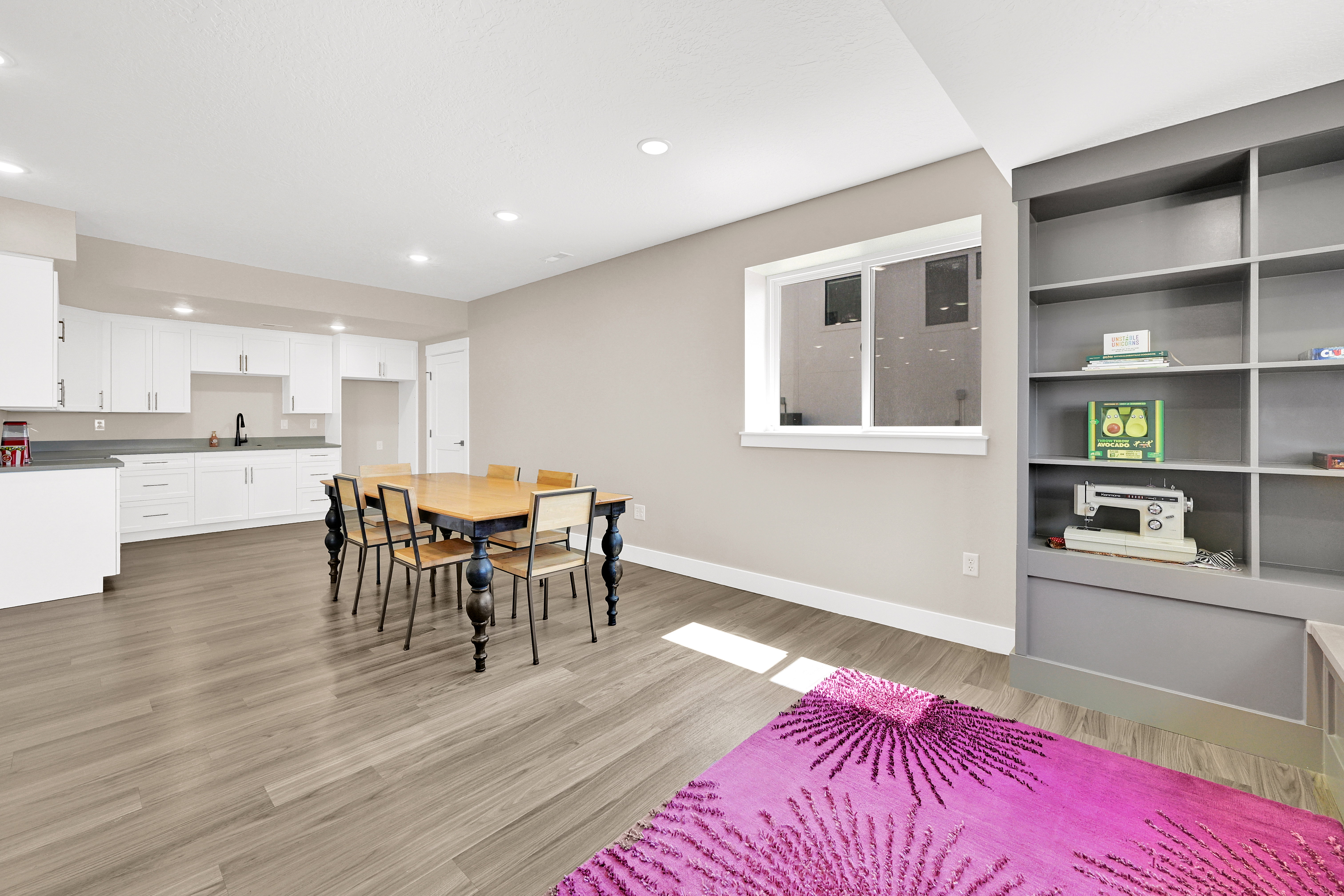 Spacious open-concept room featuring light wood-finish flooring, a full kitchen with white cabinetry and dark countertops, and a built-in shelving unit