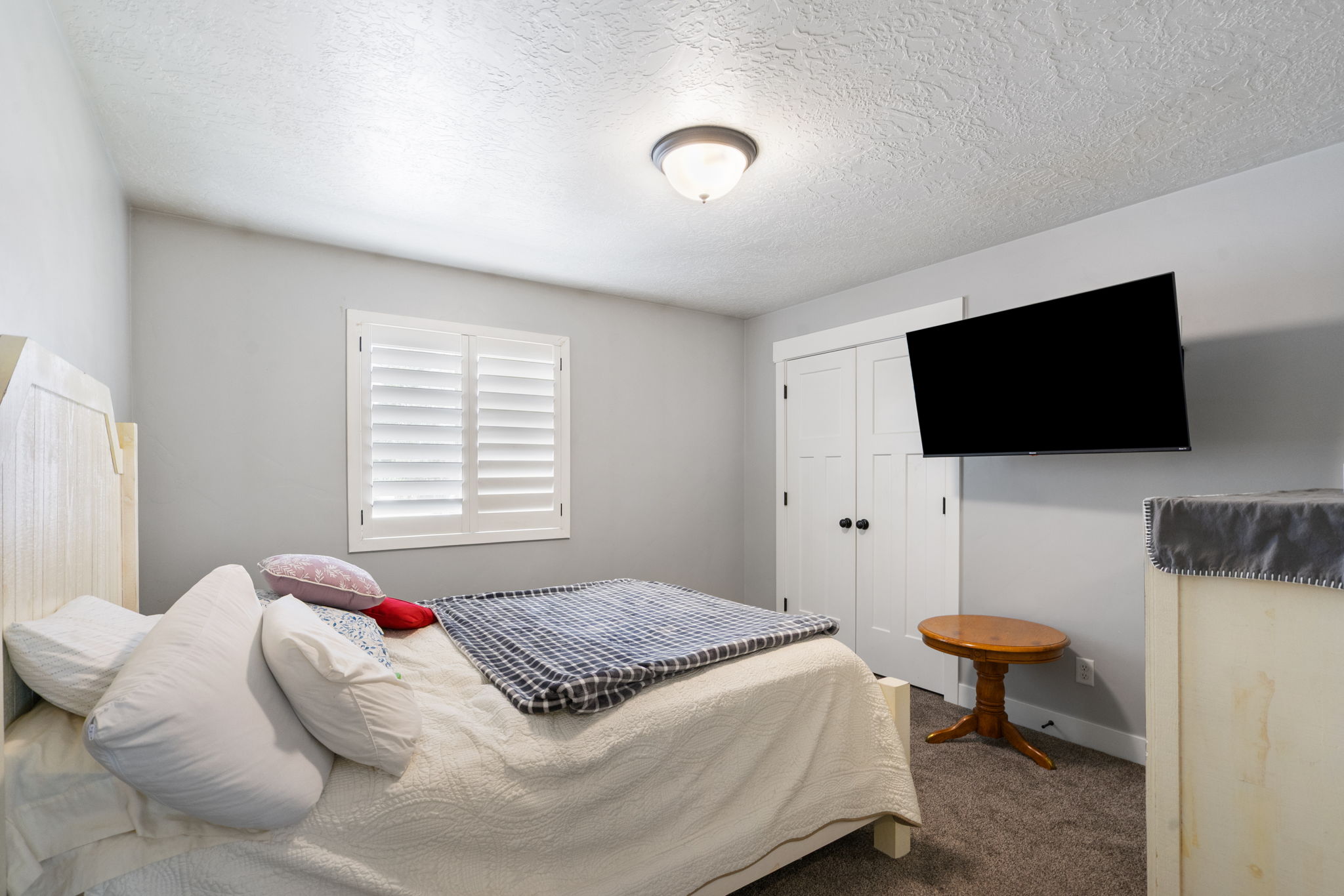 Bedroom featuring a window with white plantation shutters, neutral gray wall color, textured carpet flooring, and a ceiling-mounted light fixture