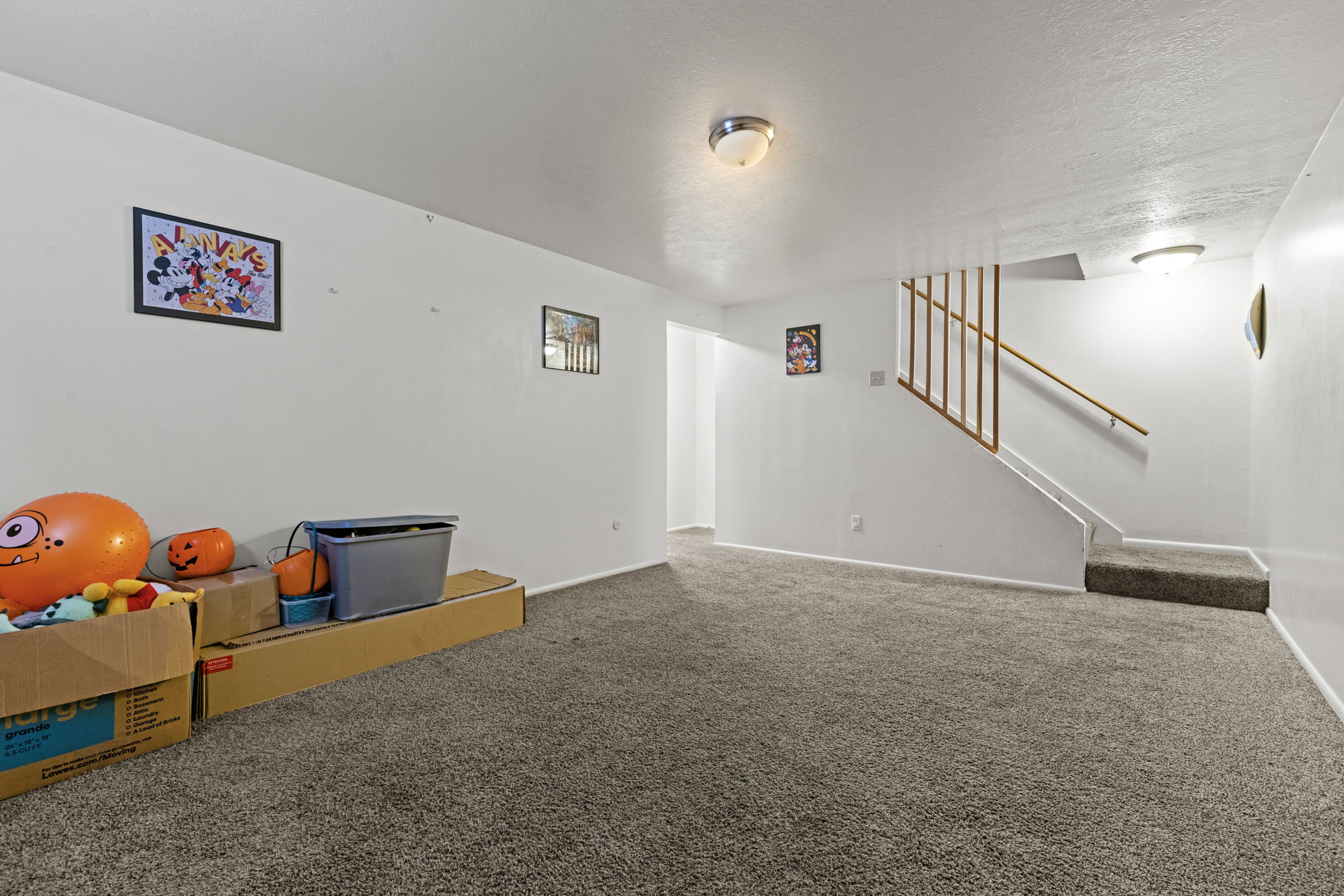 Carpeted lower-level room featuring a staircase with wood railing, white walls, and flush-mount lighting fixtures