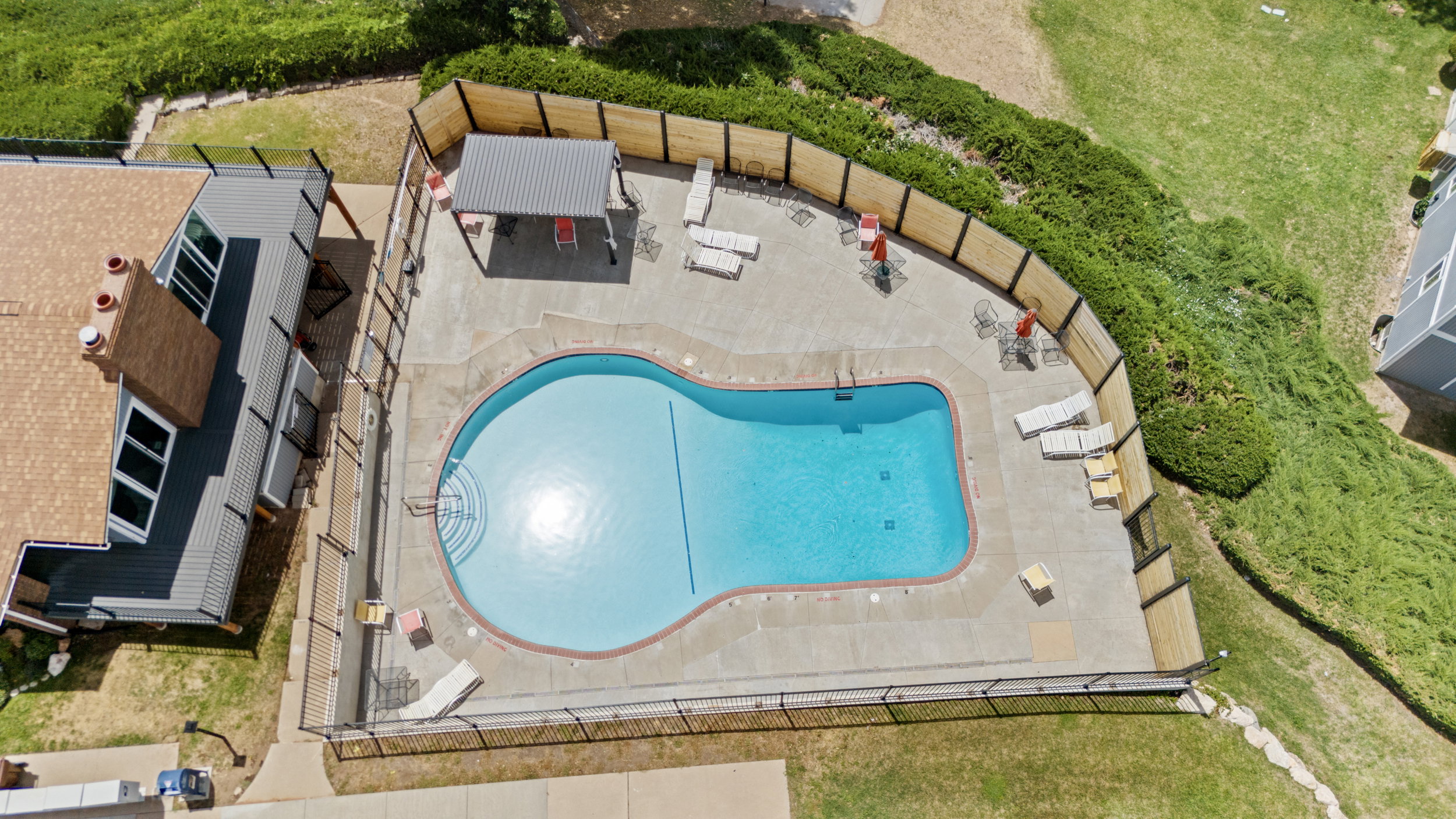 Freeform swimming pool with concrete surround, surrounded by a black metal fence and partial wooden privacy fence