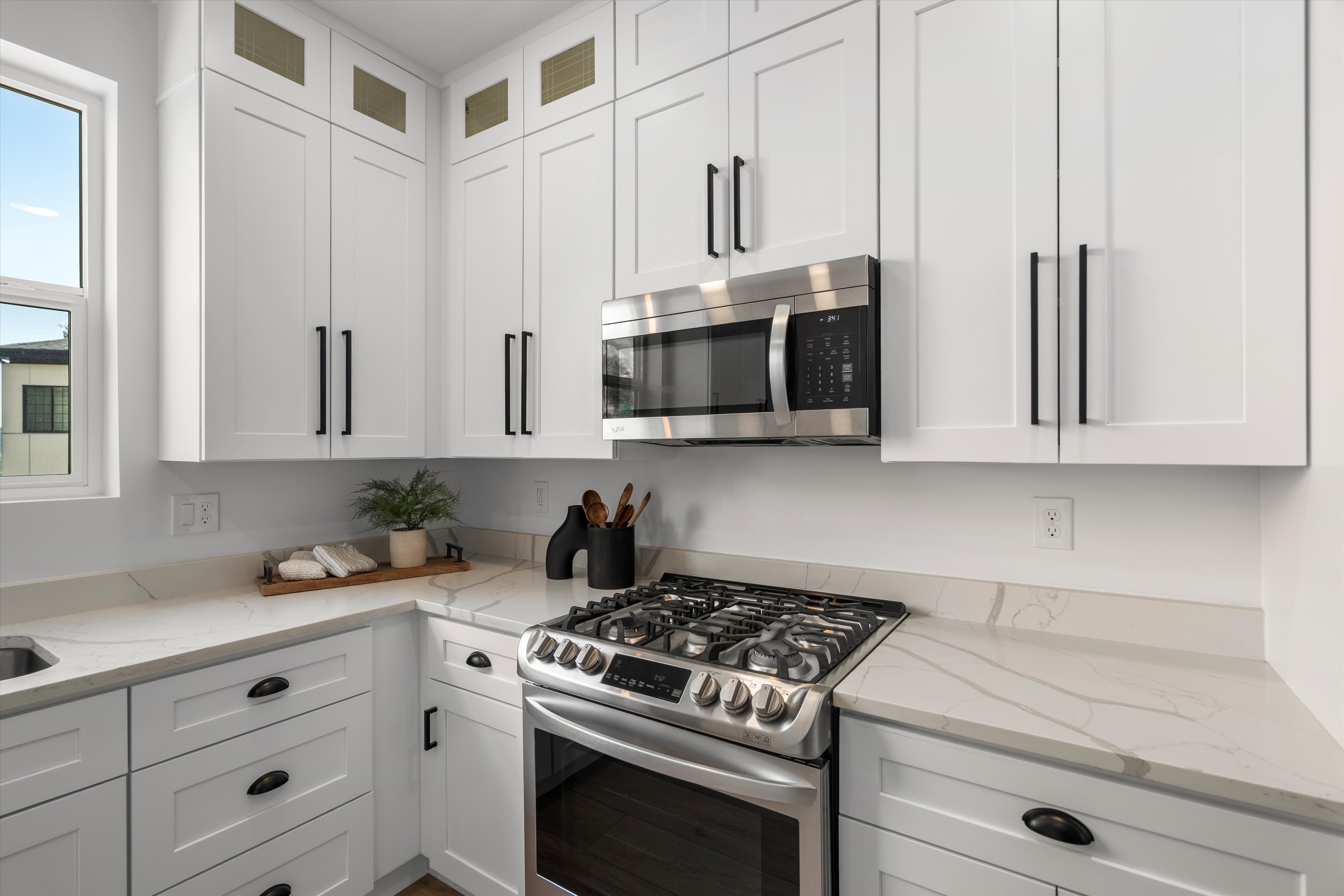 Modern kitchen featuring white shaker cabinetry with black hardware, stainless steel gas range, stainless steel microwave, and light-toned countertops with veining