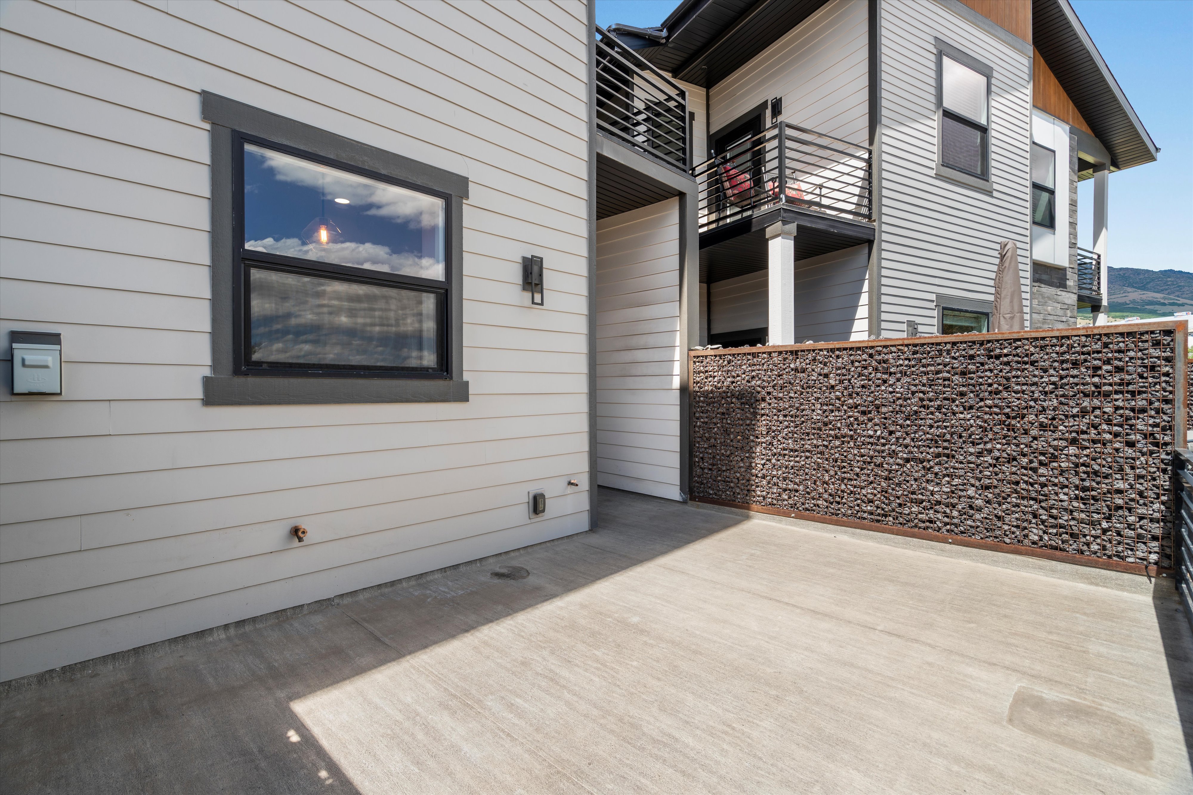 Concrete patio area featuring a modern gabion wall, light-colored horizontal siding, and a black-framed window