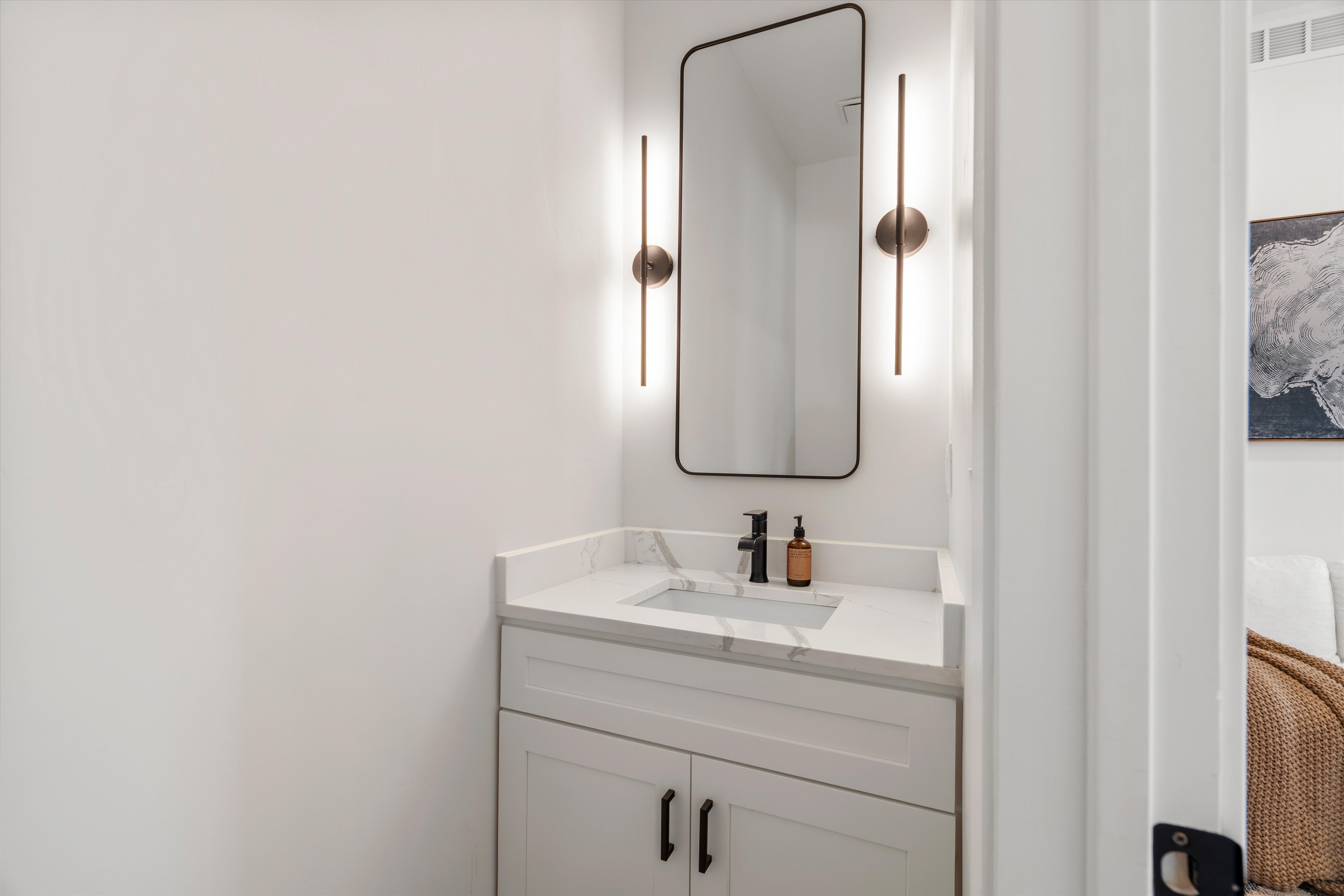 Powder room featuring a white vanity with shaker-style cabinetry, a white countertop with integrated sink, and a rectangular black-framed mirror flanked by two vertical sconces