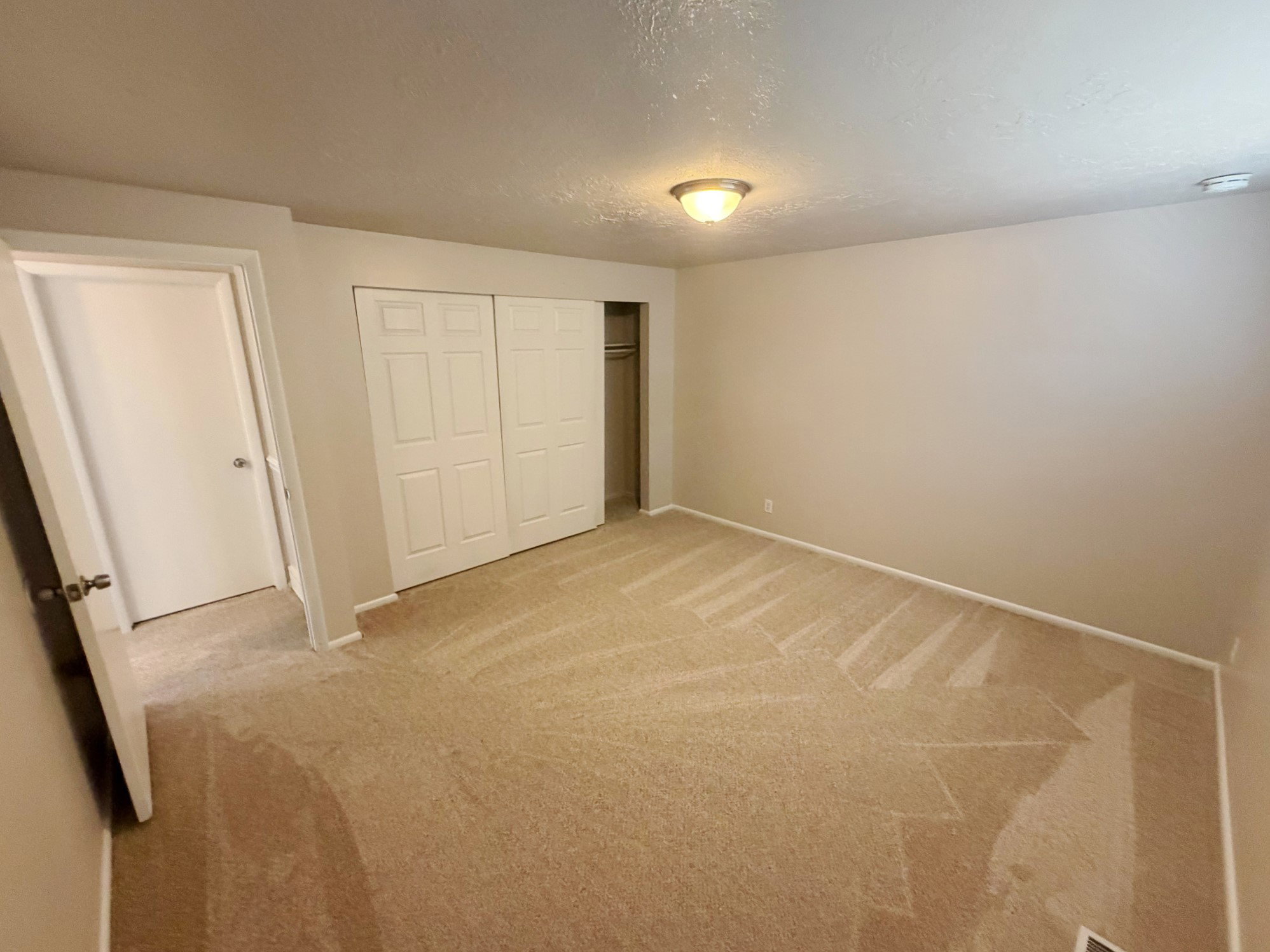 Neutral-toned walls and plush carpet flooring in an interior room featuring a ceiling-mounted light fixture, white baseboards, and a bi-fold closet with panel doors