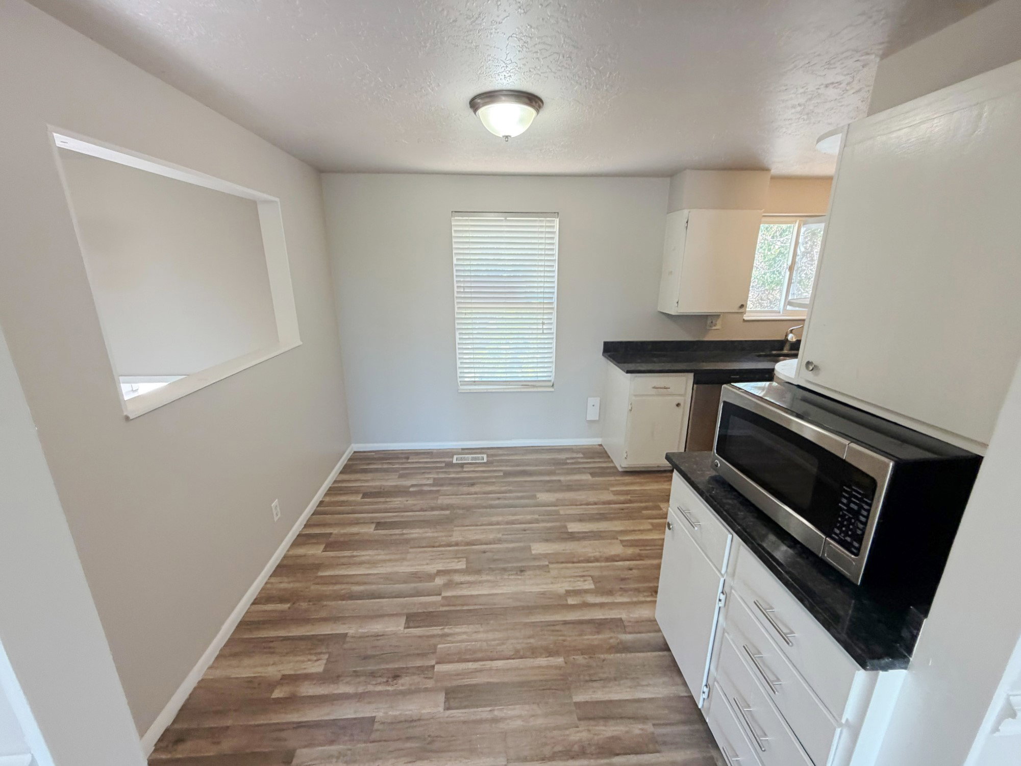 Open-concept kitchen and dining area featuring wood-finish flooring, a stainless steel microwave, white cabinetry, dark countertops, and a pass-through window