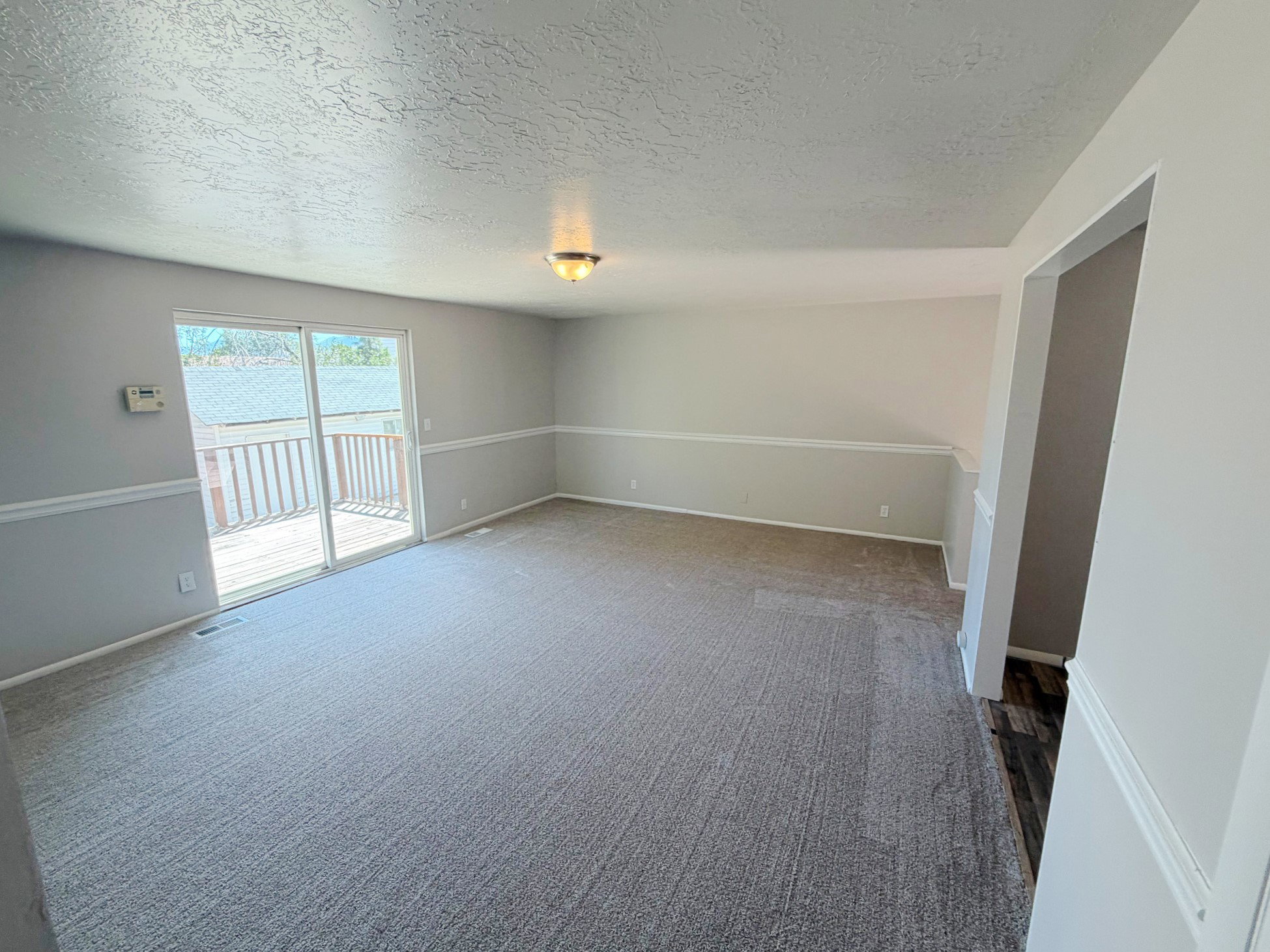 Spacious room featuring light gray walls with white chair rail molding, light gray carpeting, and sliding glass doors opening to an outdoor deck with wood railings